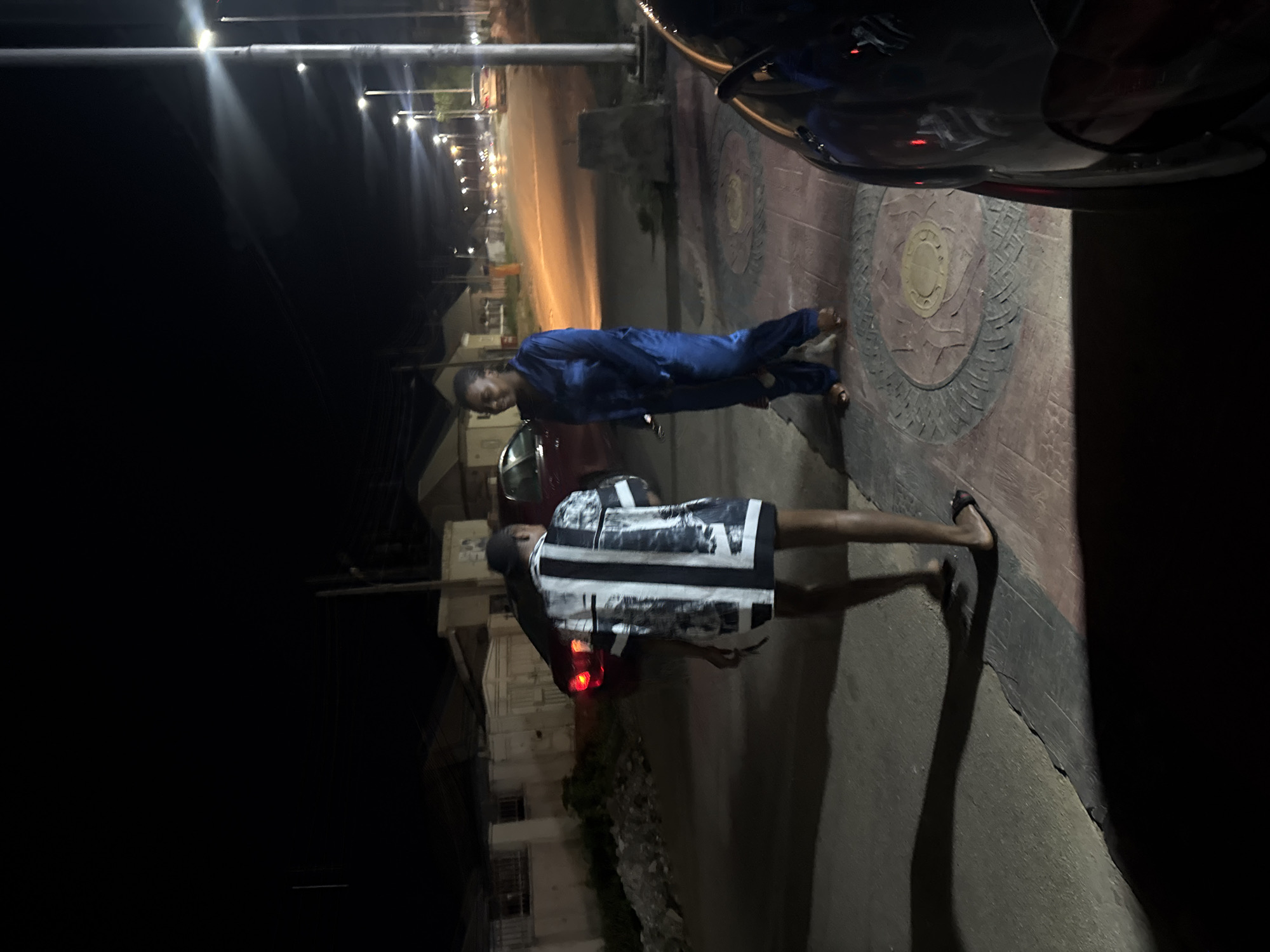 In an outdoor scene at night in Abuja, Nigeria, two individuals are visible. The person on the left, viewed from the back, is wearing a black and white patterned short-sleeved shirt and dark shorts or a skirt, and black flip-flops. They are facing away from the camera, towards the right. To their right, a second person is standing, facing left, and is dressed in a blue satin pajama set, including pants and a long-sleeved top. They are wearing brown flip-flops.
The environment is a street at night, illuminated by streetlights. The ground is paved with concrete and decorative tiles in shades of red and brown. A street curb is visible, and behind the individuals, a red car with illuminated taillights is parked. Further down the street, other vehicles and buildings are dimly visible, with streetlights lining the road, suggesting an urban or suburban setting. A concrete barrier is present near the curb. The overall lighting is dim, with harsh shadows cast by the artificial light sources.