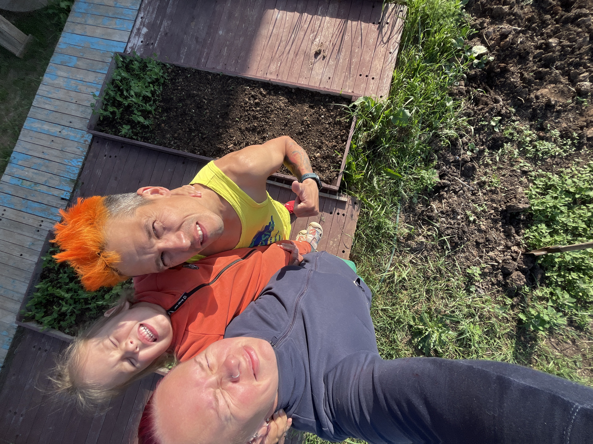 A selfie depicts three individuals outdoors, situated in a garden bed area with wooden planks and grass. In the foreground, on the left, a person with pink hair and wearing a dark blue hoodie is close to the camera, their eyes shut and their face partially obscured. To their right and slightly behind, a young child with blonde hair is nestled in the crook of another person's arm. The child is wearing an orange jacket and has their eyes squeezed shut with their mouth open, appearing to be laughing or making a face. Behind the child, a person with bright orange spiked hair and wearing a yellow tank top is looking towards the camera, with a slight smile. Their left arm is flexed, showing a tattooed bicep. The background shows a raised garden bed filled with soil and young green plants, bordered by wooden planks. The ground is covered in patches of grass and dark soil, with some visible weeds. A weathered wooden structure with blue painted sections is visible in the upper right corner. The overall scene suggests a casual, outdoor gathering, possibly in a backyard or a community garden space in Chaikovsky, Russia.