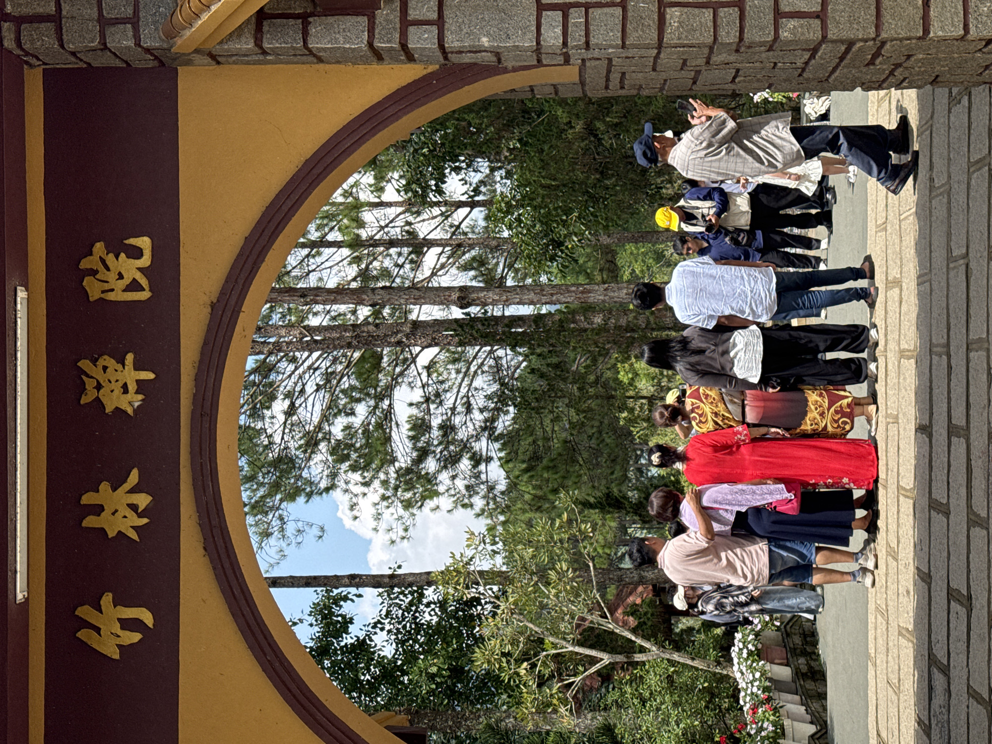 The image shows a group of people standing at the entrance of a temple, possibly the Truc Lam Zen Monastery in Da Lat, Vietnam, due to the visible architectural style and surrounding pine trees common in the region. The entrance features a large arched gateway with golden Chinese characters inscribed on a dark maroon background above.
In the foreground, the gateway is made of stone blocks and decorated with a yellow arch. A paved pathway with large, light-colored stone slabs leads towards the entrance. Beyond the arch, a lush green landscape is visible, with tall pine trees and other foliage under a bright blue sky with scattered white clouds.
A diverse group of people are gathered, some facing towards the entrance and others looking into the courtyard beyond. On the left, a man in a light pink t-shirt and denim shorts stands with his arm around a woman in a light purple shawl and dark blue skirt. To their right, a woman in a vibrant red dress and another woman in a patterned yellow and red robe are visible. Further back, a man in a blue and white striped shirt and jeans is seen, along with other individuals in casual attire. Some people appear to be taking photos or observing the surroundings.
To the right of the entrance, a man in a plaid shirt and dark pants is actively using a smartphone, holding it up as if to take a picture or video. Another man in a yellow cap and a vest is also present, possibly holding a camera. A young child in a white dress is standing near him. The overall scene suggests a visit to a place of worship or a tourist attraction.