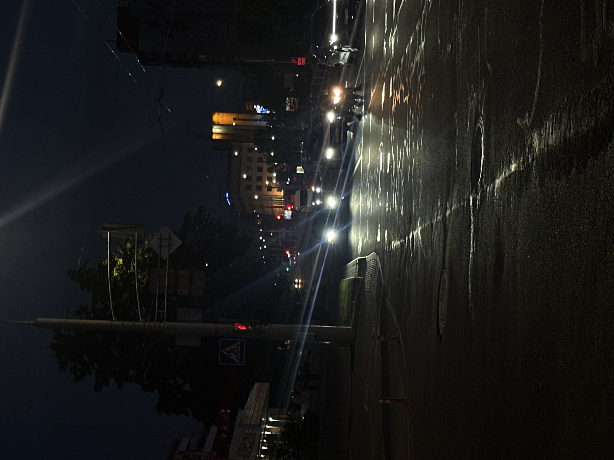 The image depicts a city street at night, possibly after rain, as indicated by the wet asphalt reflecting the lights. A traffic light is visible on the left, showing a red signal. Several cars are on the road, with their headlights on, illuminating the wet surface. Some buildings are visible in the background, some with illuminated windows and lights. A street lamp is present on the left side of the image. There are some pedestrians on the sidewalk to the left. The overall impression is of a typical urban scene at night, with active traffic.