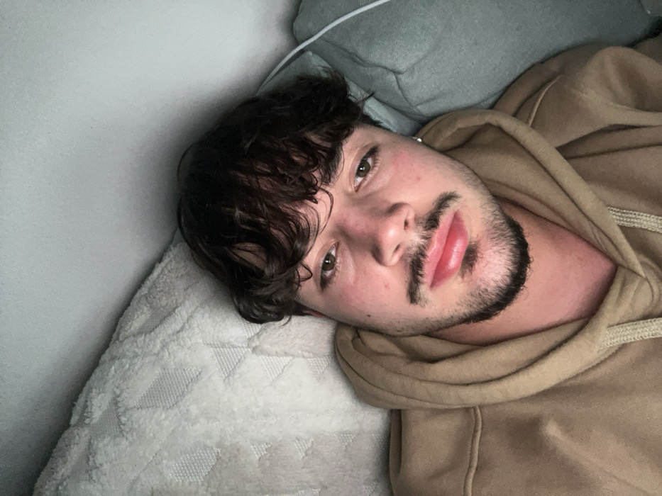 A close-up, slightly tilted image features a young man lying down indoors, looking towards the camera. He has dark, curly hair that frames his face, and a short beard and mustache. He is wearing a light brown or tan hooded sweatshirt. His eyes are open, and his expression appears neutral or relaxed, with his lips slightly parted. A small earring is visible in his left ear. He is resting his head on a light-colored pillow or cushion, and a textured white blanket or mattress cover is visible beneath him. A light grey wall or headboard is in the background. The lighting suggests an indoor setting, possibly during the daytime. No text is visible in the image.