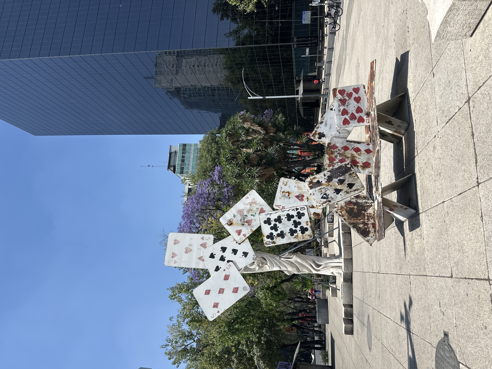 The image shows a street scene in Mexico City, Mexico. In the foreground is a metal sculpture comprised of playing cards attached to a statue, all appearing aged and weathered. The cards include symbols for hearts, clubs, and diamonds.
Behind the sculpture are trees, some bearing purple flowers, suggesting it may be spring or early summer. Further in the background are buildings, including a tall modern glass skyscraper reflecting the sky and surrounding structures. The sky is clear and blue. The scene appears to be a public area, likely a sidewalk or plaza, with pedestrians and possibly bicycles visible along the edge of the frame.