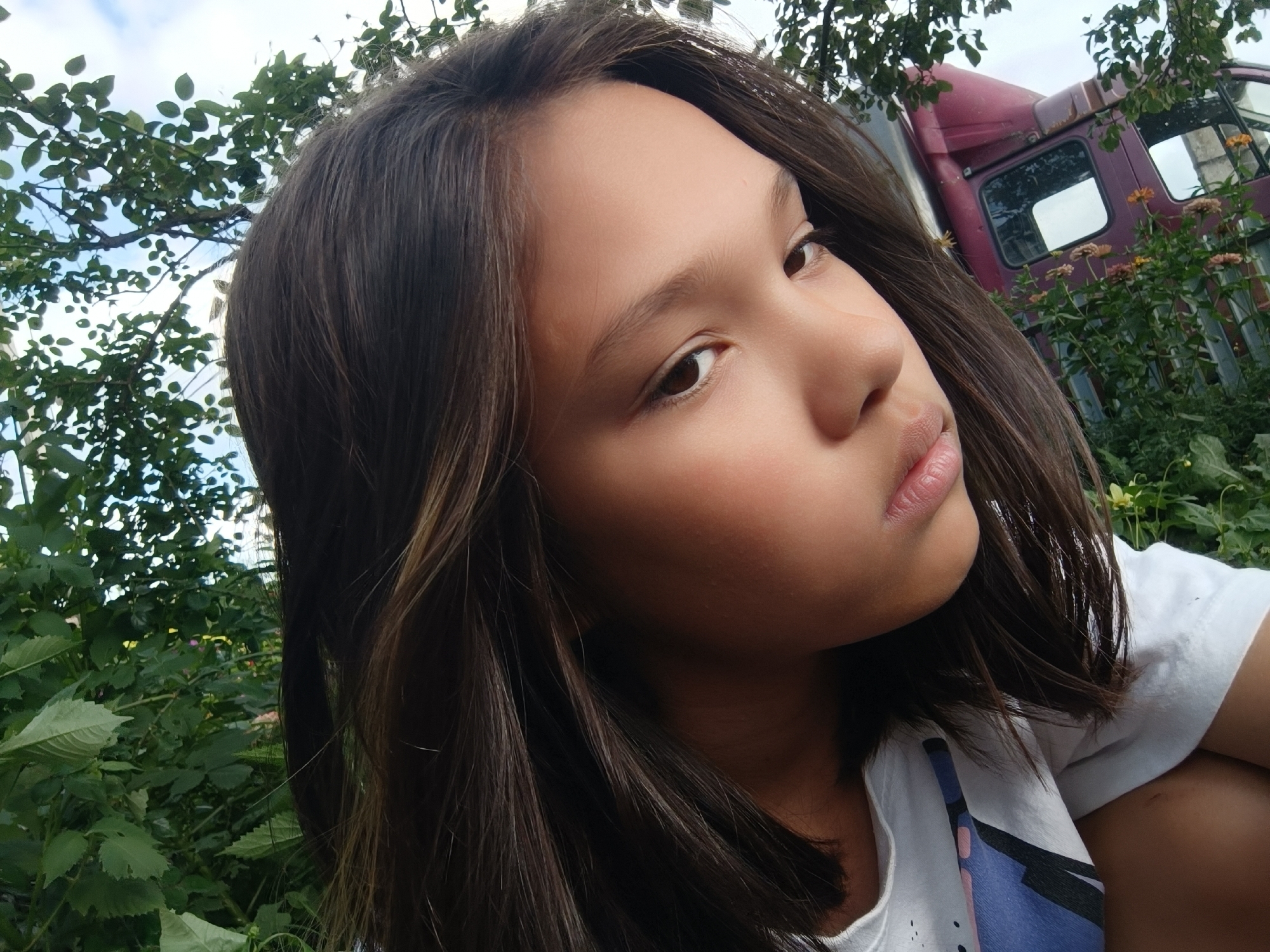 This is a selfie of a young girl with long, dark brown hair, looking off to the right of the frame. She is wearing a white t-shirt with a colorful abstract pattern on the chest. Her skin is light-toned with a warm undertone. The background is outdoors, with lush green foliage on the left and in the foreground. Behind the foliage on the right, a portion of a weathered, dark red truck is visible, along with some flowering plants with pale orange and pink blossoms. The sky is visible in the upper left corner, appearing overcast. The lighting suggests it is daytime. The overall mood is candid and relaxed, with the girl's gaze directed away from the camera. There is no visible text in the image. The setting suggests a rural or semi-rural environment, consistent with the provided location context of Karsha, Russia.