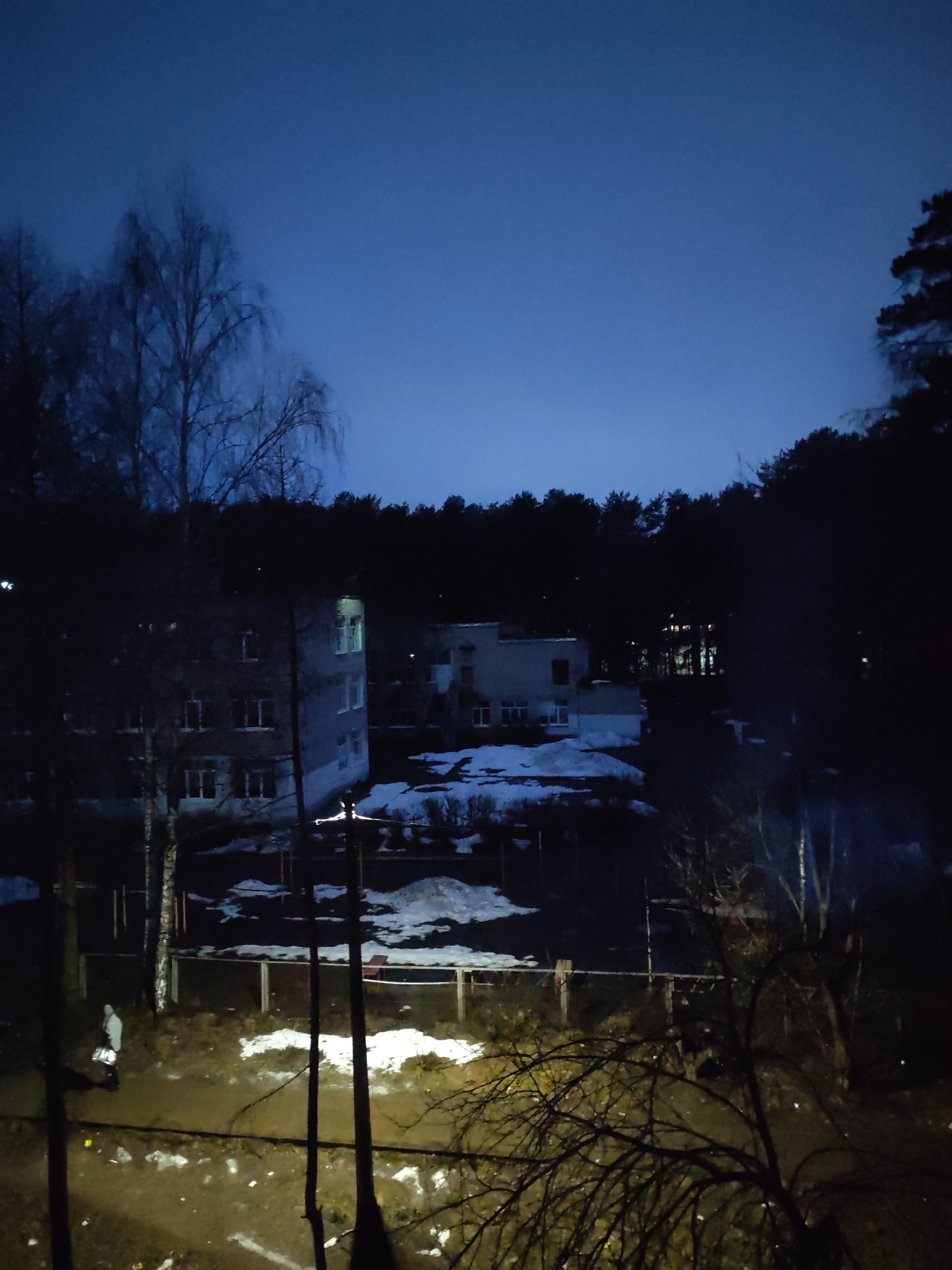 This nocturnal image captures an outdoor scene at dusk or night, likely in a semi-urban or suburban area bordering a forest. The sky is a deep blue, not completely dark, indicating twilight.
In the foreground and mid-ground, patches of melting snow are visible amidst dirt and mud, suggesting late winter or early spring conditions. A sparse, dark wooden fence runs horizontally across the mid-ground. Tall, bare deciduous trees, possibly birches, stand prominently on the left, with their intricate branches silhouetted against the sky. A dense line of dark coniferous trees forms a forest in the background and on the right.
Two multi-story buildings are seen in the middle distance. The one on the left is greyish with multiple windows, some of which are illuminated, casting warm light. Another lighter-colored building is further back on the right. Additional lights are visible deeper within the forested area, hinting at more structures or distant streetlights.
A solitary person is visible in the lower left quadrant, seen from behind. They wear a light-colored hooded jacket or coat and appear to be carrying a white object, possibly a bag, which brightly reflects light from an unseen source. This light source, or possibly a streetlight, illuminates the ground around the person.
The overall atmosphere is quiet and somewhat desolate due to the low light and the transitional seasonal elements. No visible text is present in the image. The scene is located in Chaikovsky, Russia.