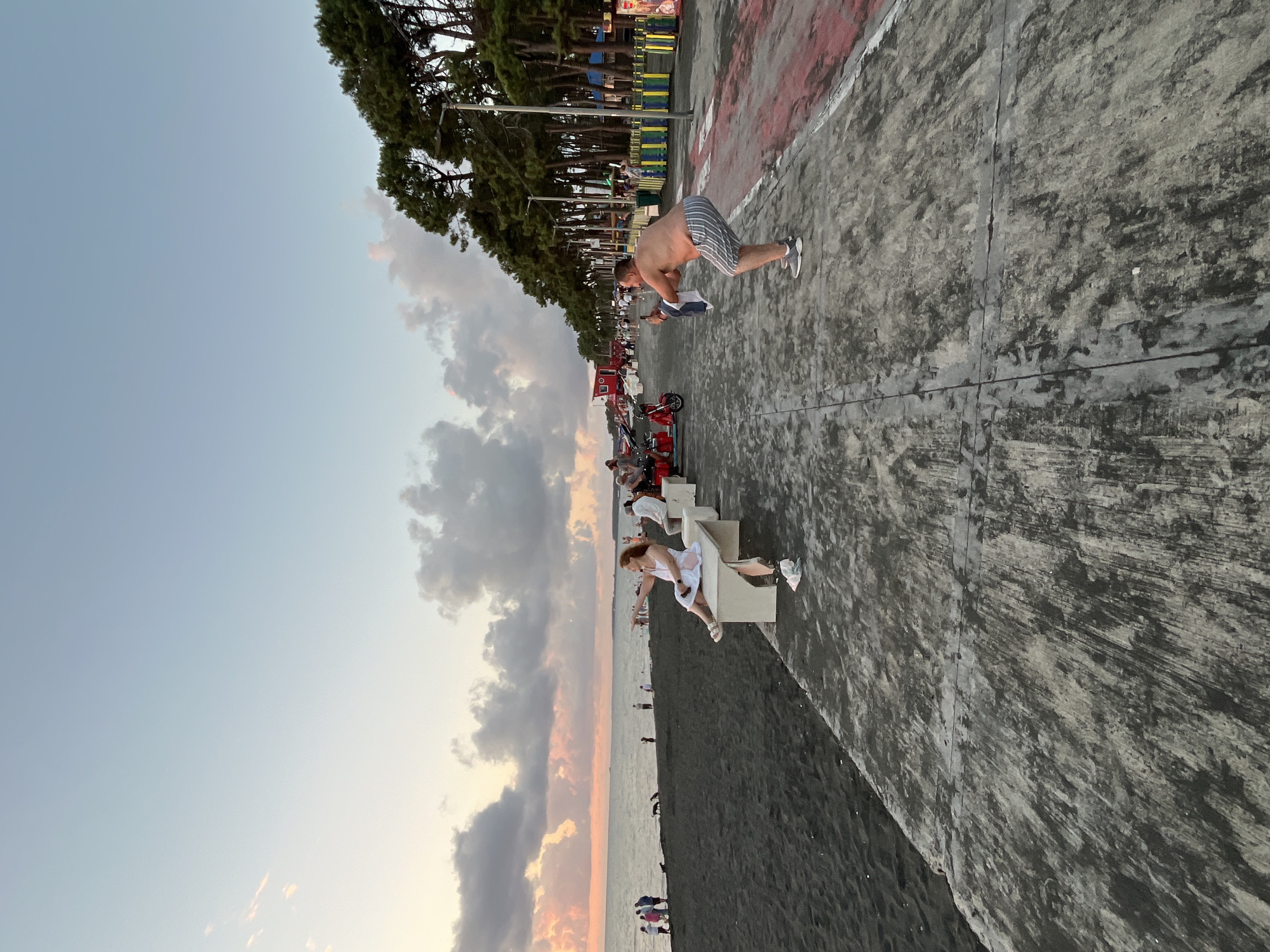 The image depicts a scene on the Ureki, Georgia beach promenade at dusk. The promenade is paved with concrete and has red and white markings on the right side, suggesting a designated path. A line of tall pine trees borders the promenade on the right, with streetlights visible among them. On the left side of the promenade is a black sand beach adjacent to the sea, where the sky is a pale blue with scattered clouds tinged with pink and orange from the sunset.
Several people are present. In the foreground, a man with striped shorts is crouching to take a photo with his phone, holding a white towel in his left hand. Further down the promenade, a woman in a white dress is seated on a bench, pointing towards the sea. Other people are seen walking and sitting on benches, and some are on the beach or near red scooters. The overall atmosphere appears relaxed, characteristic of a seaside resort area.