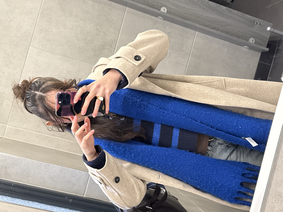 A woman in a tan coat takes a mirror selfie. She wears a blue, fringed scarf over a striped sweater and jeans. Her hair is in a messy bun. She holds a smartphone with a multicolored case in front of her face. Her fingers display several rings. The background includes tiled walls and a glimpse of a purse.