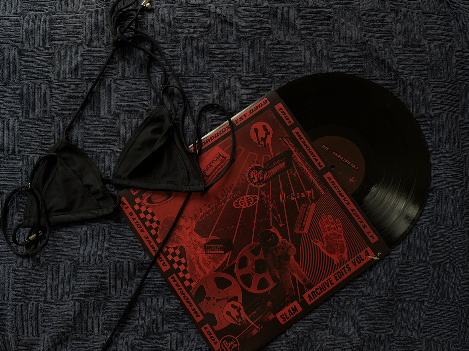 The scene is located in Budva, Montenegro. A black two-piece bikini top, consisting of two triangular fabric cups with thin tie straps, lies on a dark blue, woven, textured surface. Small golden-colored metallic beads are visible at the ends of some straps. Partially overlapping and situated beneath the bikini top is a 12-inch vinyl record sleeve. The sleeve features a black background with red graphical elements, including stylized film reels, abstract machinery, a checkered pattern, and a depiction of a hand. Text visible on the sleeve includes "SLAM," "ARCHIVE EDITS VOL. 4," "1001 MEMORIAM," and "RESIST." A black vinyl record is partially extracted from the sleeve, extending towards the bottom right of the frame. No individuals or active interactions are present.