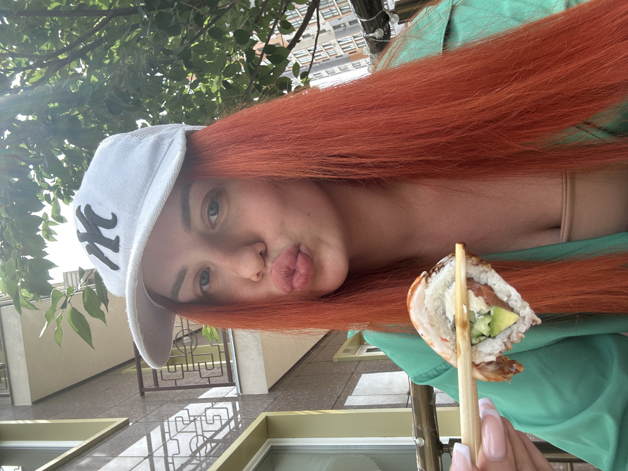 A woman with long, bright orange hair, wearing a white baseball cap with a black "NY" logo and a teal jacket, is holding a piece of sushi with chopsticks. She is making a kissing face at the camera. The background shows a building with a balcony and trees. The location is identified as Lymanka, Ukraine.