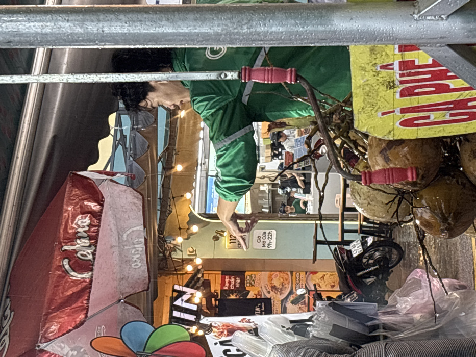The image captures a street-level scene in Ngũ Hành Sơn Ward, Vietnam, under artificial lighting, suggesting dusk or night. In the immediate right foreground, a male individual wearing a green Grab delivery uniform with reflective strips and a 'G' logo is seen from the torso up, looking left and pointing his right index finger forward. His hair is dark and curly. A wet, metallic pole runs vertically along the extreme right edge of the frame. Below the Grab rider, a cluster of wet, brown coconuts is visible, with a red-handled tool embedded among them. Partially obscured on the left side of the coconuts is a yellow sign with red Vietnamese text, displaying "CÀ PHÊ" (Coffee).
Behind the Grab rider and slightly to the left, a storefront with a light green facade is visible. A white sign affixed to the facade displays the number '112' and the text "Giờ Mở Cửa 7h-22h" (Opening Hours 7 AM - 10 PM). Inside the brightly lit establishment, a counter area is visible with at least two individuals: one female in a green shirt behind the counter and another person in a light-colored top further back. Dark wooden tables and stools are also present inside. Above the storefront, a red awning, marked with the faded white script "Calana", extends over the street, with a string of bare-bulb lights suspended underneath. To the far left, a colorful pinwheel (red, orange, green, blue) is attached to a vertical element, alongside stacks of clear plastic cups and plastic bags. A dark-colored motorcycle, displaying a license plate "43-LD 078.XX", is parked on the street to the left of the storefront entrance. Posters advertising "Ramen chuẩn Nhật" (Japanese standard Ramen) are visible on the wall adjacent to the motorcycle.