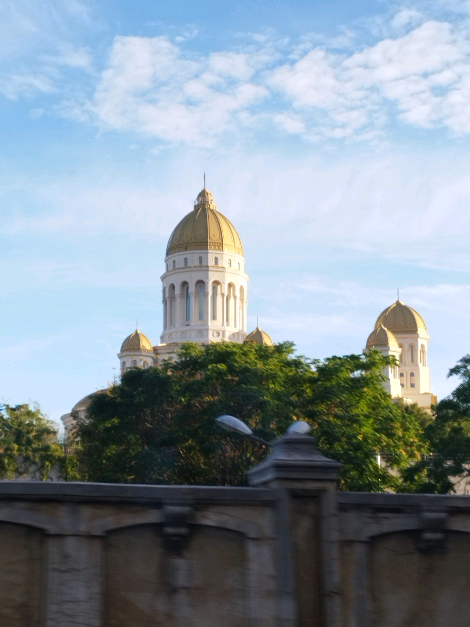 The image depicts the Romanian Orthodox Cathedral in Bucharest, Romania, viewed from a low angle with a bright blue sky and scattered white clouds overhead. The main facade of the cathedral, with its prominent golden domes, is visible behind a lush green canopy of trees. In the foreground, a blurred, gray stone structure, possibly a wall or bridge railing, partially obscures the view of the cathedral. The overall scene is bathed in natural light, suggesting it was taken during the daytime.