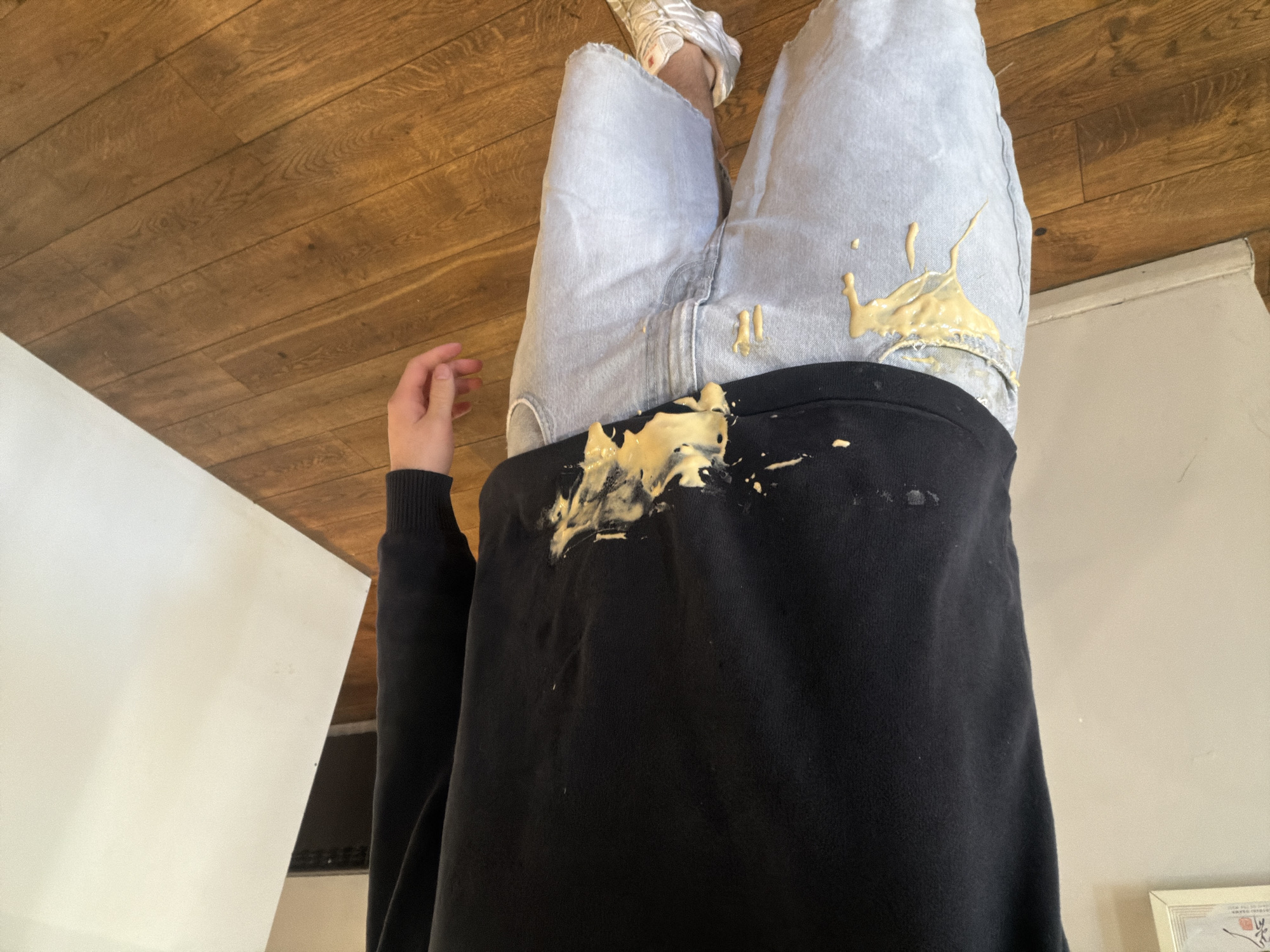 The scene, located indoors in Sofia, Bulgaria, depicts an individual from the chest down, standing on a light brown wooden plank floor. The person is wearing a black long-sleeved sweatshirt and light blue denim shorts. Both garments are extensively soiled with a thick, viscous, yellow-beige substance, consistent with a food product. This substance is splattered across the lower front of the sweatshirt and drips onto the upper left thigh and pocket area of the denim shorts. Additional small, darker stains are present on the upper portion of the black sweatshirt. The individual's right hand is visible, hanging naturally at their side. A white athletic shoe with a thick sole and orange accents is visible on the right foot. The background features plain light-colored walls, with a framed item on the left wall and a large white rectangular panel leaning against the right wall, partially obscuring a dark object.