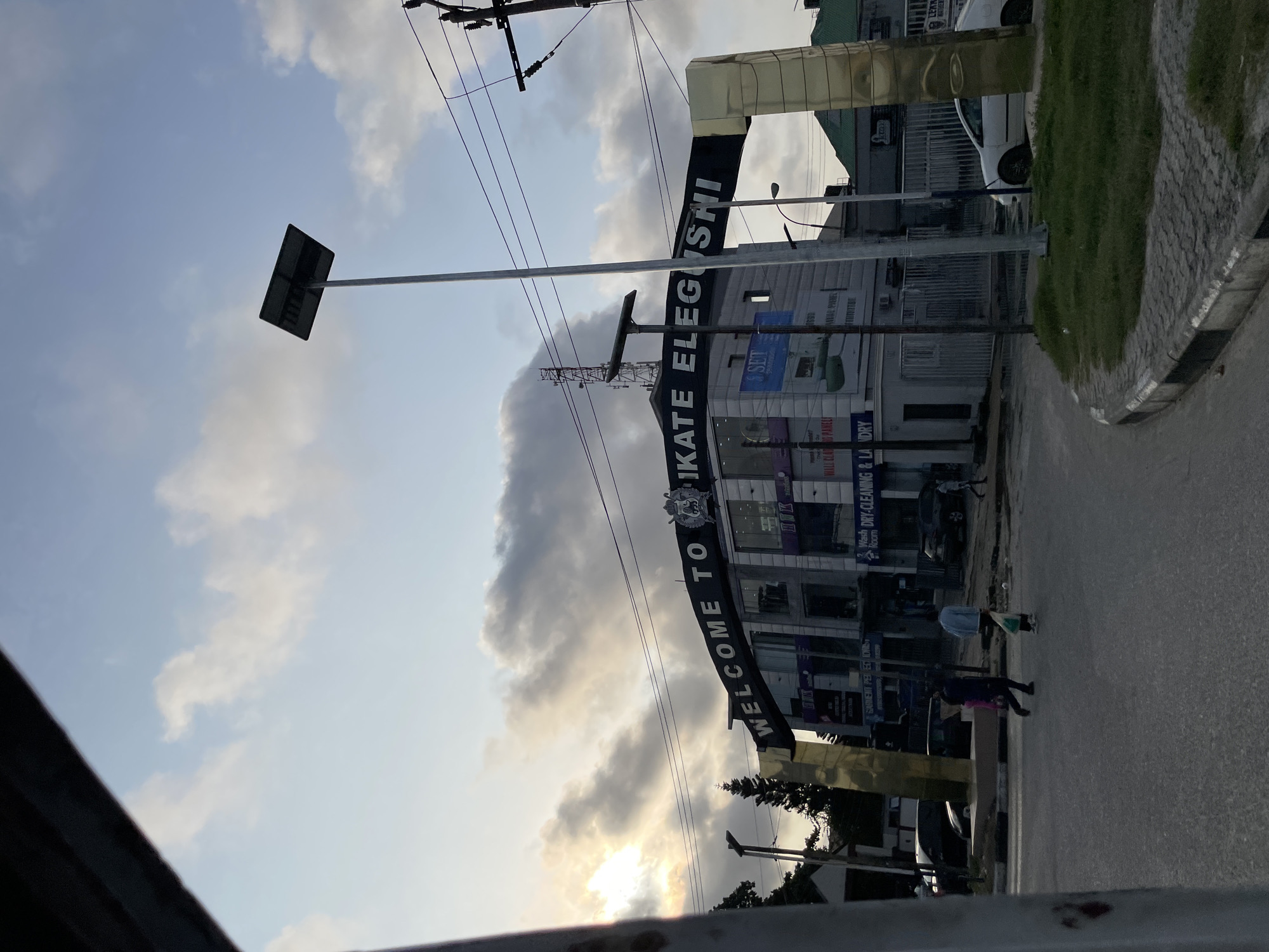 The image shows an urban street scene in Alaguntan, Nigeria, under a cloudy sky at dusk. A large archway spanning the road displays the text "WELCOME TO IKATE ELEGUSHI." The archway is supported by golden pillars and is positioned in front of a multi-story building with various businesses visible, including "SET Prestige Dry Cleaning & Laundry" and "Garment Perfections." Several vehicles are present on the street, including cars and what appears to be a bus or large van. Pedestrians are visible crossing the street. A street lamp with a solar panel is mounted on a pole near the archway, and utility poles with electrical wires are also present. The road surface is asphalt, with a paved walkway and a grassy area alongside. The overall impression is a busy commercial area in a Nigerian city.