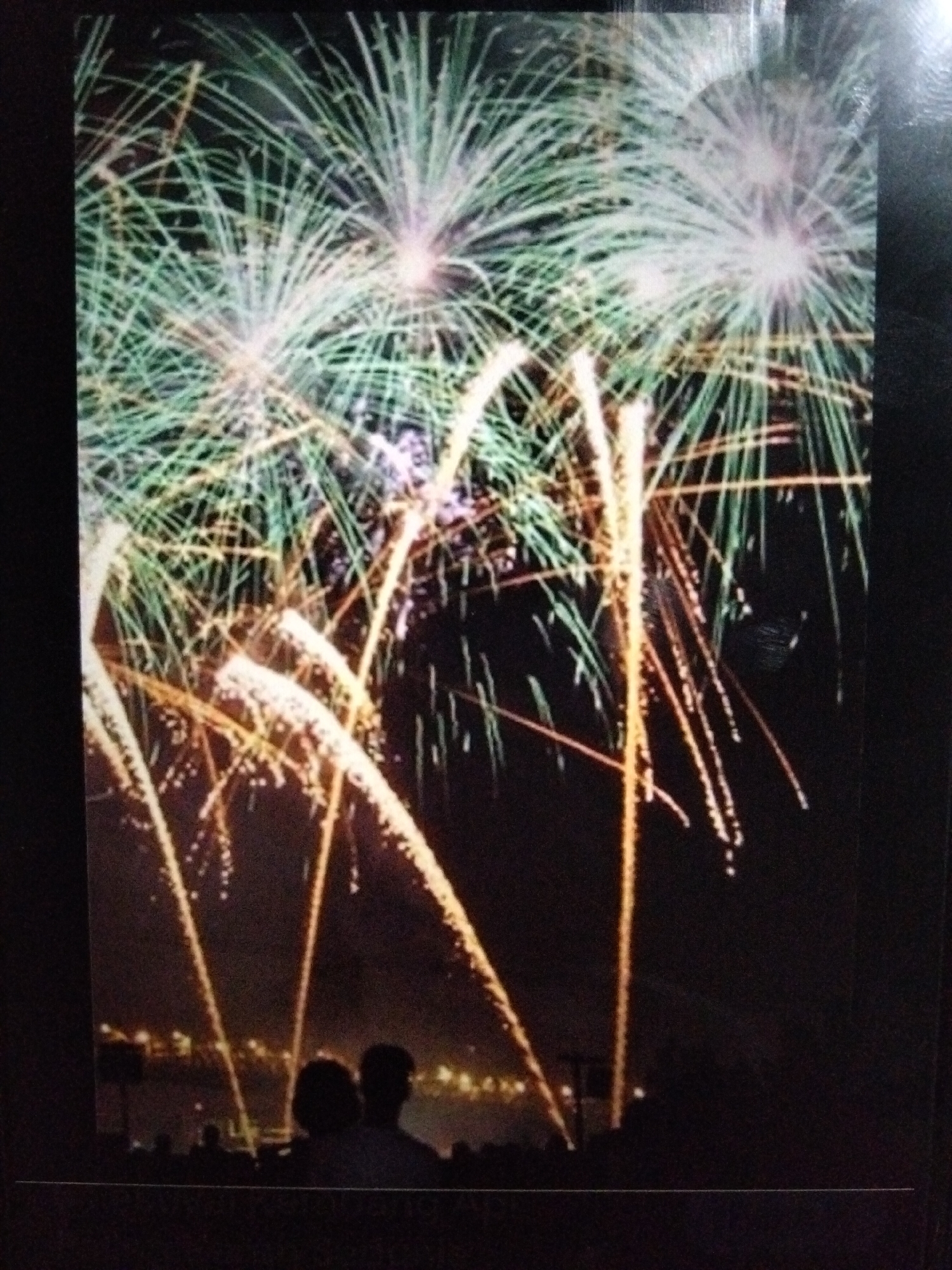 The image depicts a nighttime scene featuring fireworks illuminating the sky over a cityscape. Several large, fan-shaped fireworks with green and white light streaks dominate the upper portion of the frame. Below these, orange, glowing trails of light descend from bursts of fireworks, some appearing as thin lines and others as wider streaks with smaller dots of light. In the foreground, silhouetted figures of people are visible, appearing to watch the fireworks display. Two figures in the middle ground are more defined, with one appearing to have longer hair. A faint outline of buildings and lights from the city can be seen in the distance, beneath the fireworks. The overall environment is dark, indicating the display is taking place at night. There is no specific indication of the location being Kualaraja, Indonesia, within the image itself.