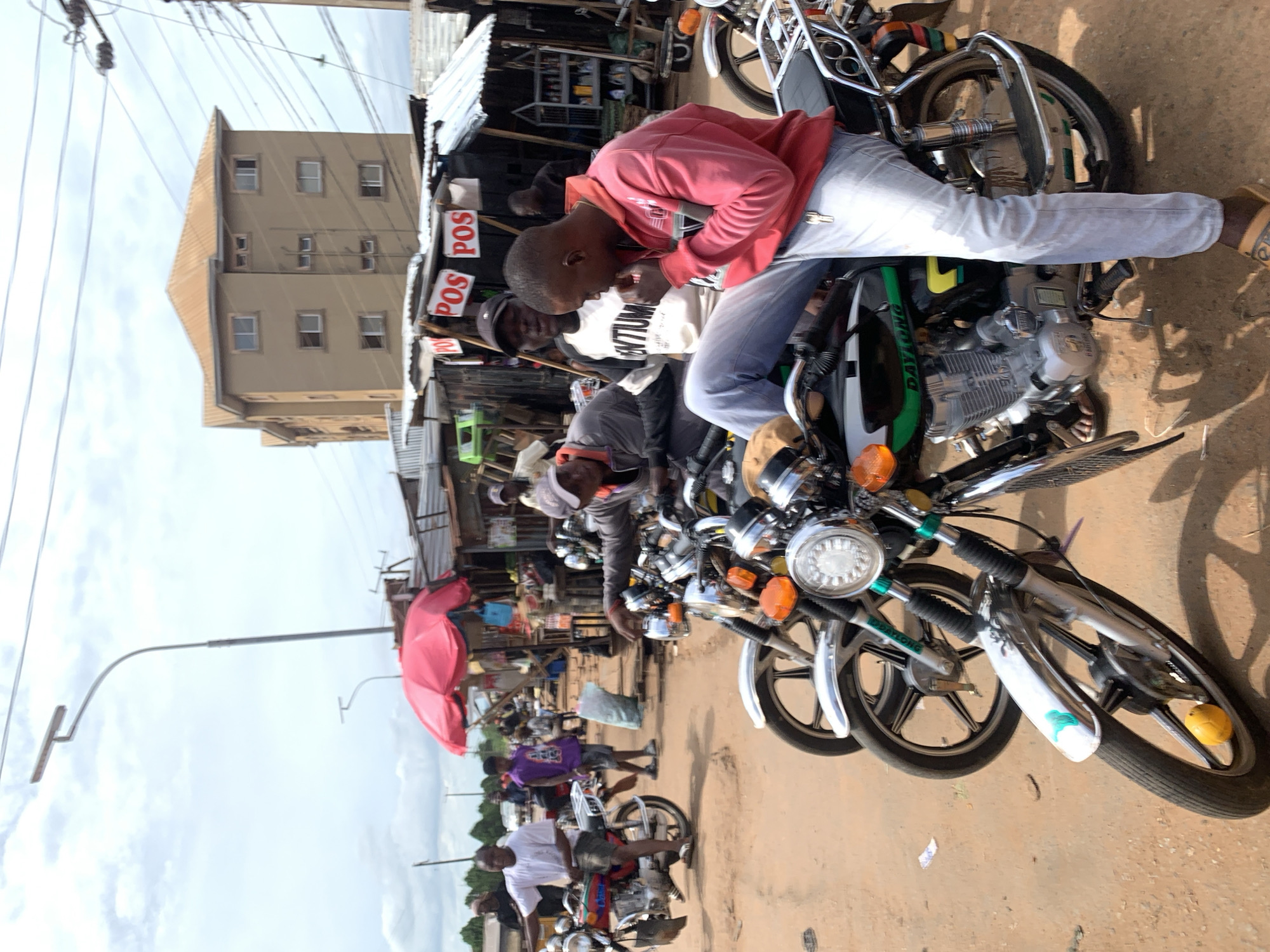 The image depicts a street scene in Nsugbe, Nigeria. Several motorcycles are parked along the side of a dirt road. A young man in a red hoodie and light-colored jeans is sitting on one of the motorcycles. Other individuals are visible near the motorcycles. A building with multiple stories is visible in the background. The street is lined with stalls and vendors under makeshift corrugated iron roofs. A red umbrella is open. A street lamp extends over the road. Power lines run overhead.
