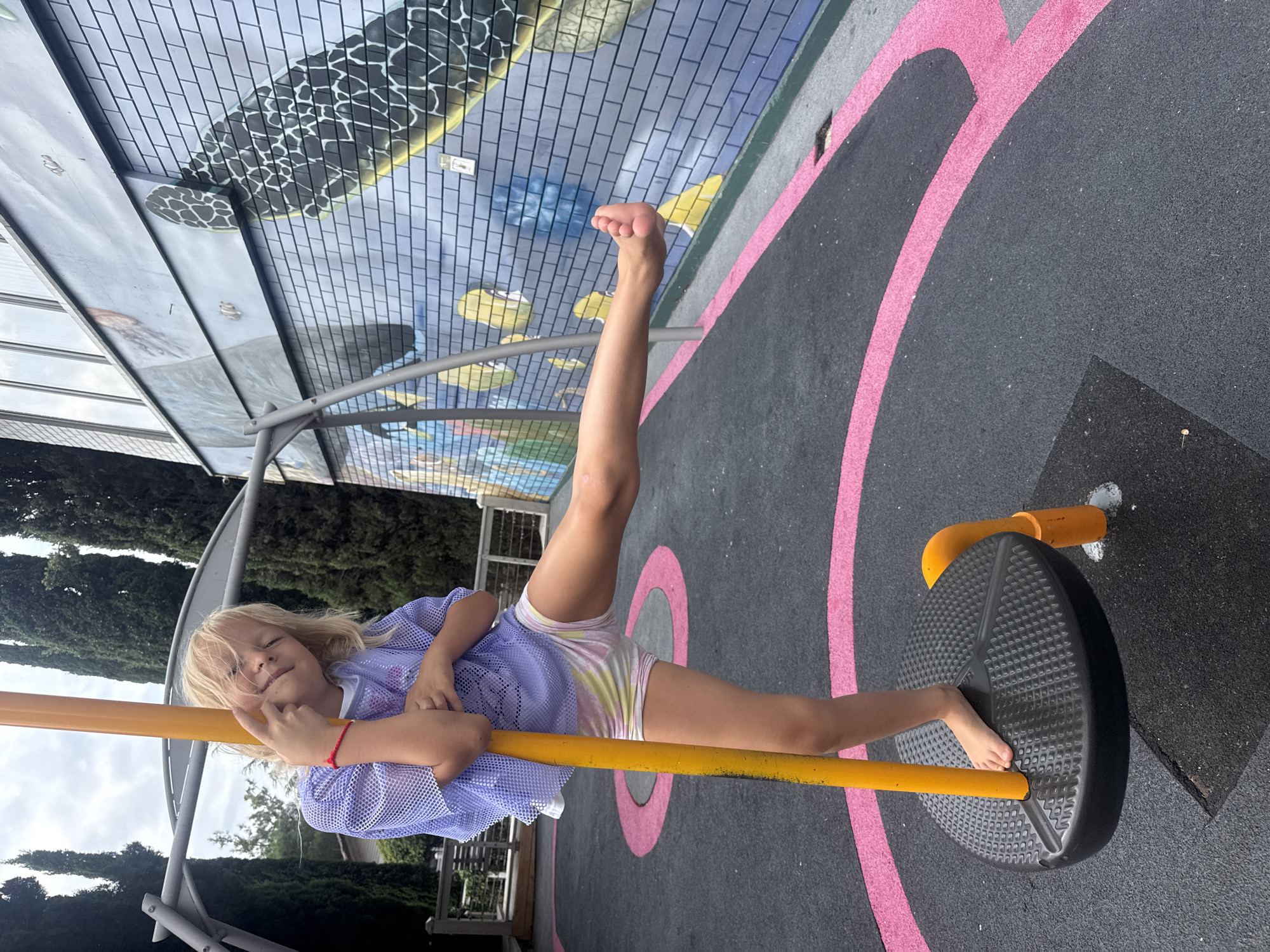 A young girl with blonde hair is posing on a playground spinner. She is wearing a light purple mesh top and tie-dye shorts. Her left foot is on the black spinner platform, and her right leg is extended outward. She is holding onto a yellow pole. The playground surface is black rubber with pink circular markings. In the background, there are tall trees and a building with a mural on its side depicting a turtle and other aquatic life. The scene appears to be an outdoor playground.
