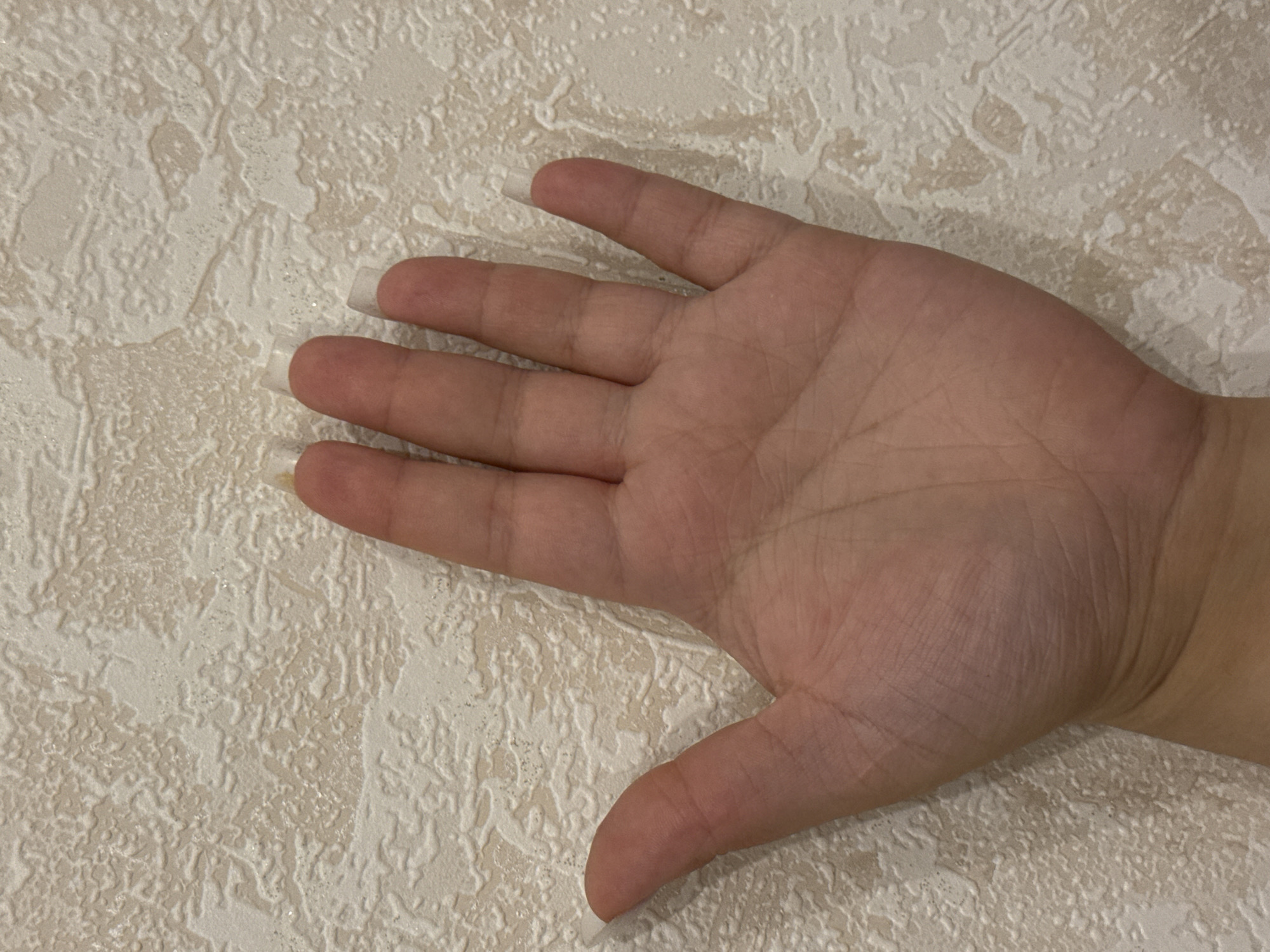 A person's left hand is shown held up against a textured, light-colored wall. The hand is positioned palm-out, with fingers slightly spread. The fingers have long, square-shaped artificial nails. The background is a light beige wall with a raised, uneven texture that appears to be wallpaper. No specific location in Saratov, Russia can be determined from the image.