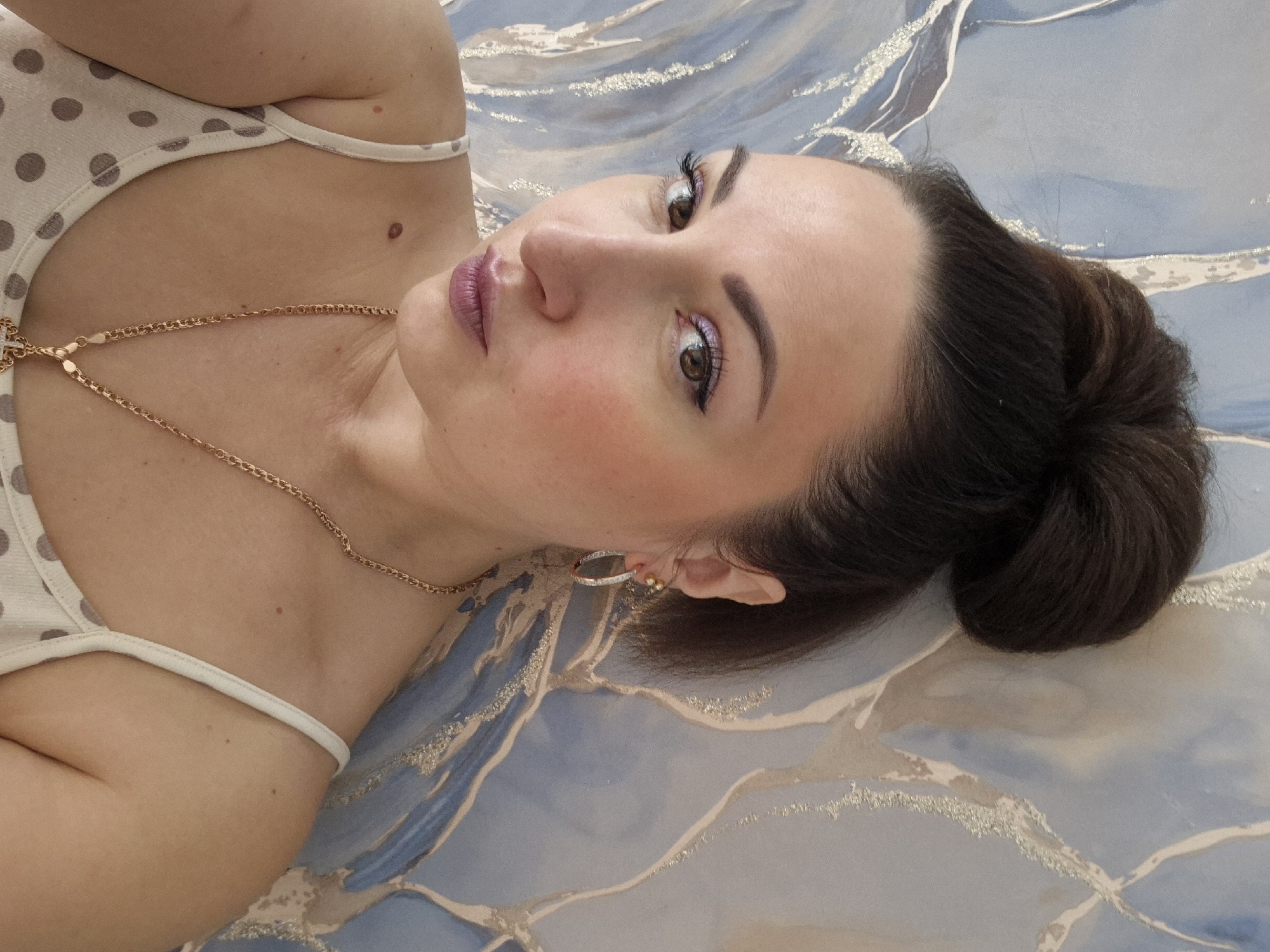 A woman with brown hair pulled back in a bun is lying on a surface with a blue and silver marbled pattern. She is wearing a cream-colored tank top with brown polka dots and a thin gold necklace. She is looking directly at the camera. Her skin tone is fair, and she is wearing makeup, including blush and eye shadow. She has hoop earrings in her ears. There is a mole visible on her chest. The location is in the city of Unknown, Unknown.
