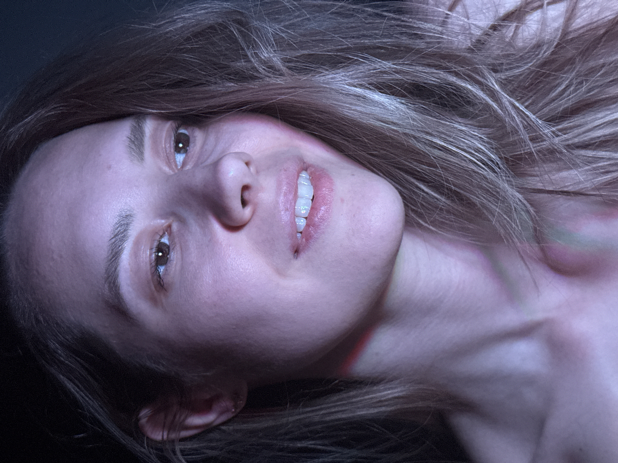 The image presents a close-up, tilted view of a fair-skinned woman with brown hair. She is smiling, and her head is positioned at an angle, suggesting she may be lying down. There are red and green streaks on her neck, possibly indicative of markings or discoloration. The environment is dark and undefined, with no clear indication of the location being Chaikovsky, Russia, or any other specific place. No specific objects or interactions are discernible beyond the woman herself.