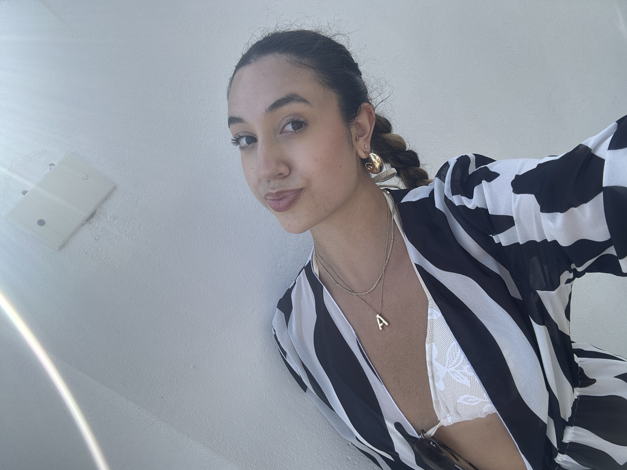 A woman is pictured in a selfie taken outdoors against a textured white wall. She is wearing a black and white patterned garment over a white lace bikini top. She has on a gold necklace with an "A" pendant, and large gold hoop earrings. Her hair is pulled back in a braid. Sunlight beams across the top left of the image, and a white fixture is visible on the wall. The image appears to have been taken in San Juan, Puerto Rico.
