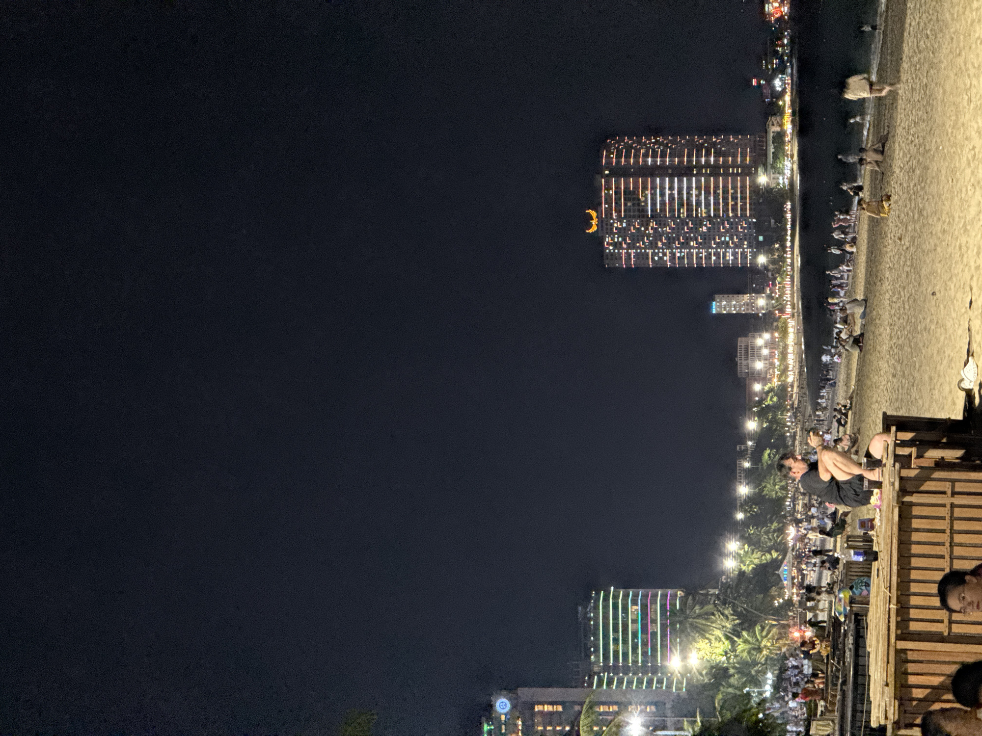 The image captures a nighttime view of Nha Trang, Vietnam, showing a bustling beach scene alongside illuminated city buildings. The beach is crowded with people, some walking along the shore, others sitting and interacting, and some near bamboo structures, possibly part of beachside establishments. A person is seen sitting on one of the bamboo structures, looking at the beach. Palm trees and other foliage are interspersed along the beach area.
In the background, several buildings are visible, adorned with decorative lighting. One distinctive building has brightly colored vertical lights, while others exhibit grid-like window patterns with yellow lighting. The sky is dark, contrasting with the illuminated cityscape and beach below.