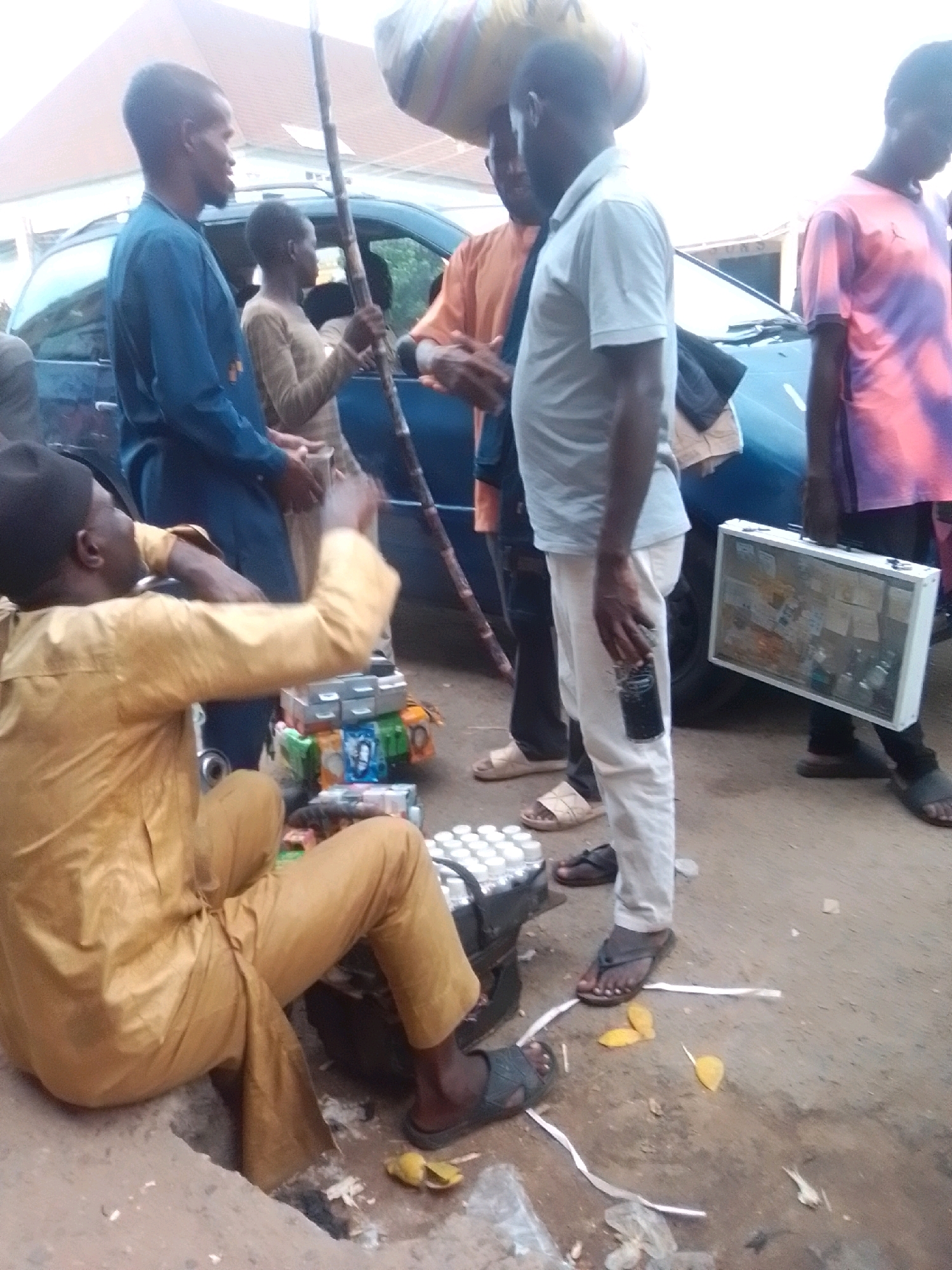 The image shows a street scene in Arawa, Nigeria, with several individuals present. In the foreground, a man wearing a yellow traditional outfit and a black cap is seated, appearing to be attending to a display of goods. Next to him are several boxes and stacks of bottled drinks. To his right, another man in a light-colored polo shirt and white trousers stands holding a black object, possibly a phone or a small device, in his right hand. He is also holding a white briefcase-like container filled with various items. Behind him, a person in a blue shirt is standing near a dark-colored car. Further to the right, a person wearing a pink and purple tie-dye t-shirt is visible. In the background, a building with a red roof is partially visible. The ground is paved and appears dusty. The overall scene depicts a bustling outdoor environment, likely a marketplace or roadside stall.
