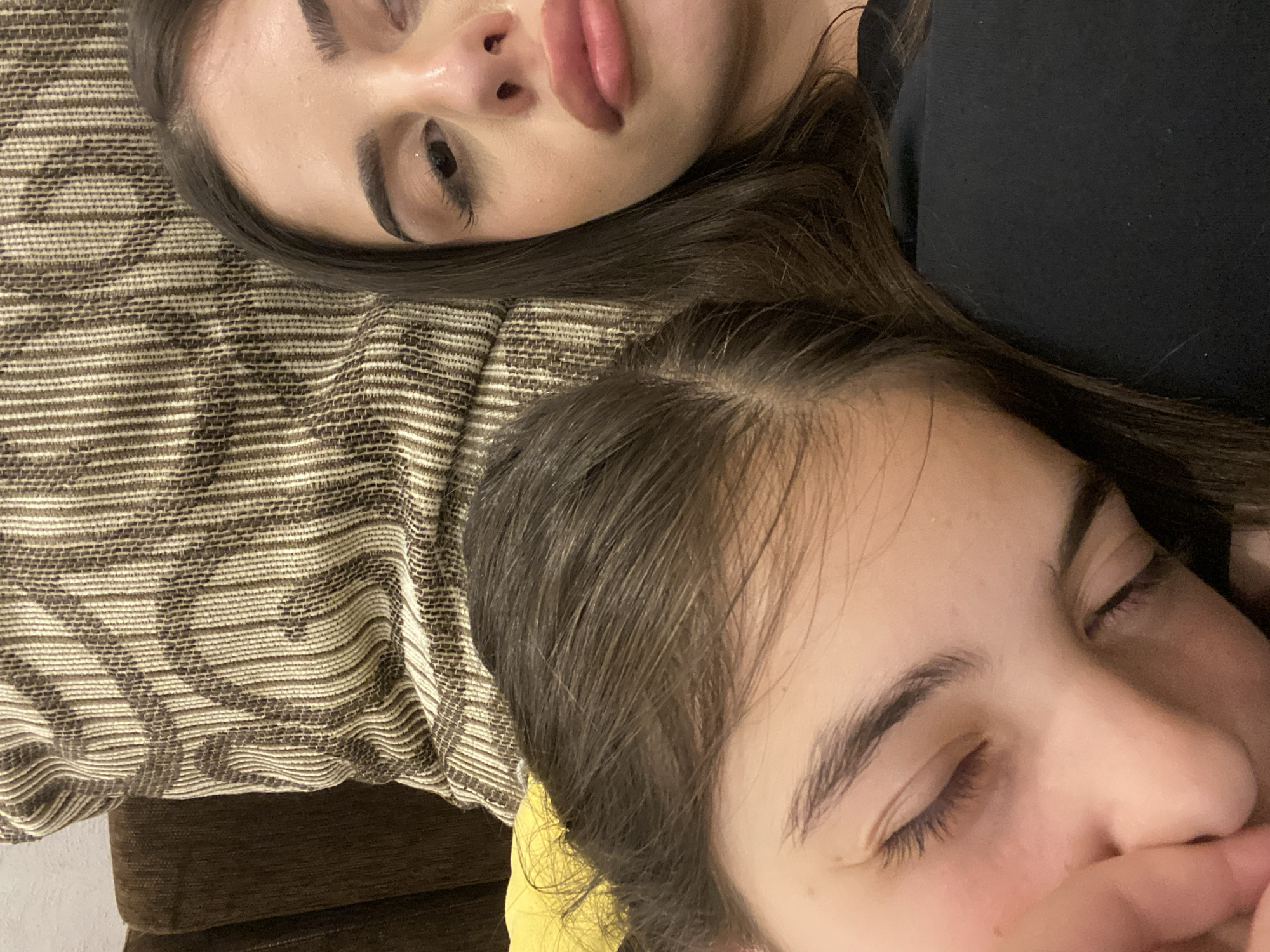 Two young women are lying down close to each other. The woman on top is looking up with her eyes open, and the other woman has her eyes closed. They are lying on a striped cushion. The location is unknown and not identifiable in the image.
