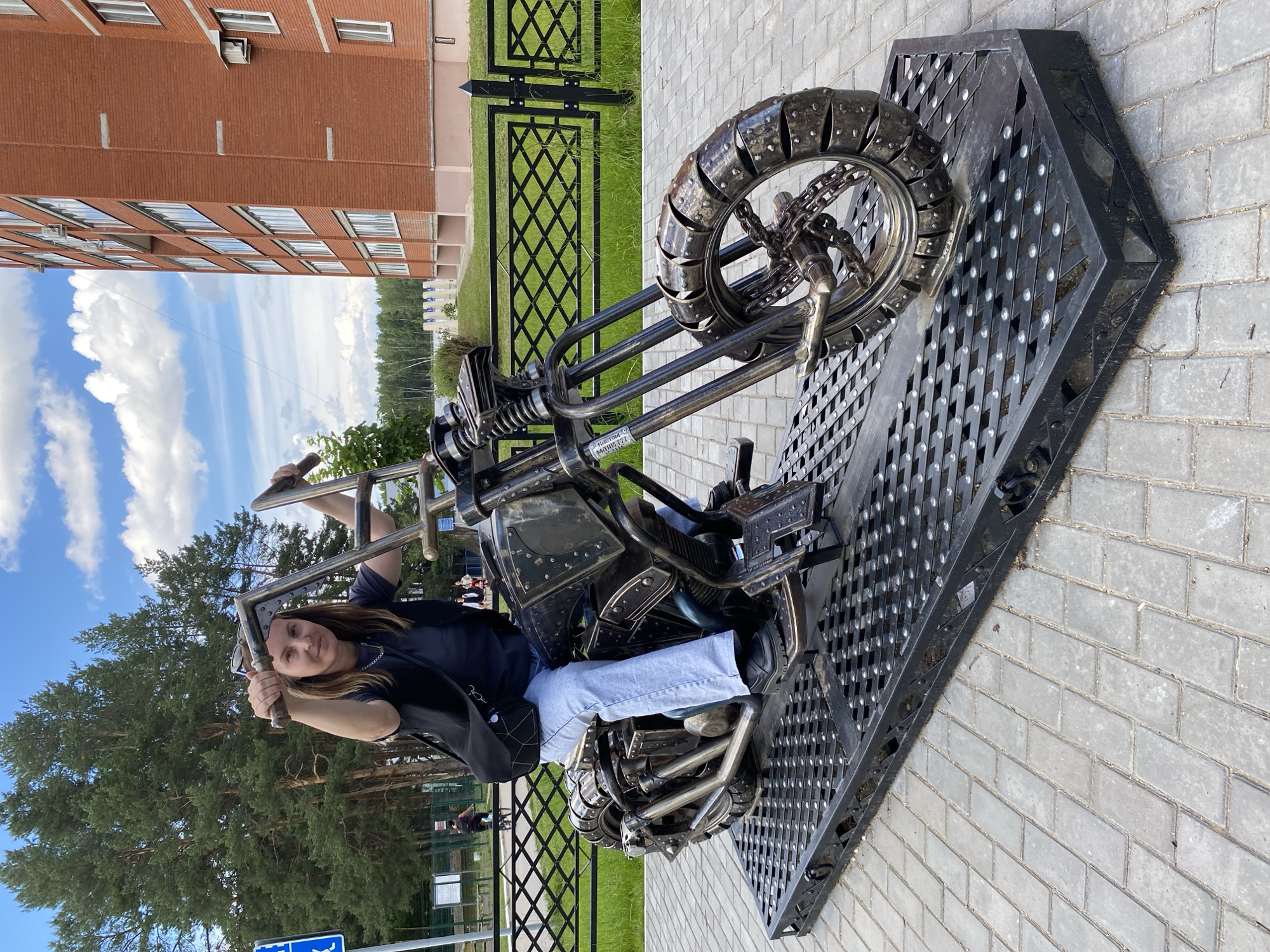 A woman is sitting on a decorative metal motorcycle sculpture outdoors. The motorcycle is dark colored with intricate metalwork, including a detailed wheel and engine components. It is mounted on a dark metal platform with a diamond-patterned surface. The woman is wearing a dark shirt, blue jeans, and black shoes. She is holding onto the handlebars and looking towards the camera with a relaxed expression.
The setting appears to be a public outdoor space. Behind the motorcycle, there is a black metal fence with a diamond pattern, and beyond that, a grassy area. Further in the background, there is a tall brick building with many windows, and a line of trees. The sky is visible with some clouds. The image is taken from a slightly elevated angle.
There is some visible text on the motorcycle sculpture, on a small plate near the seat, which appears to be a manufacturer's mark or model number. It reads "Изготовлен-777" (Made-777).
The lighting suggests it is daytime, likely mid-morning or afternoon, with bright sunlight. The overall impression is of a casual, pleasant day spent outdoors, possibly in a park or town square. The location context is Chaikovsky, Russia.