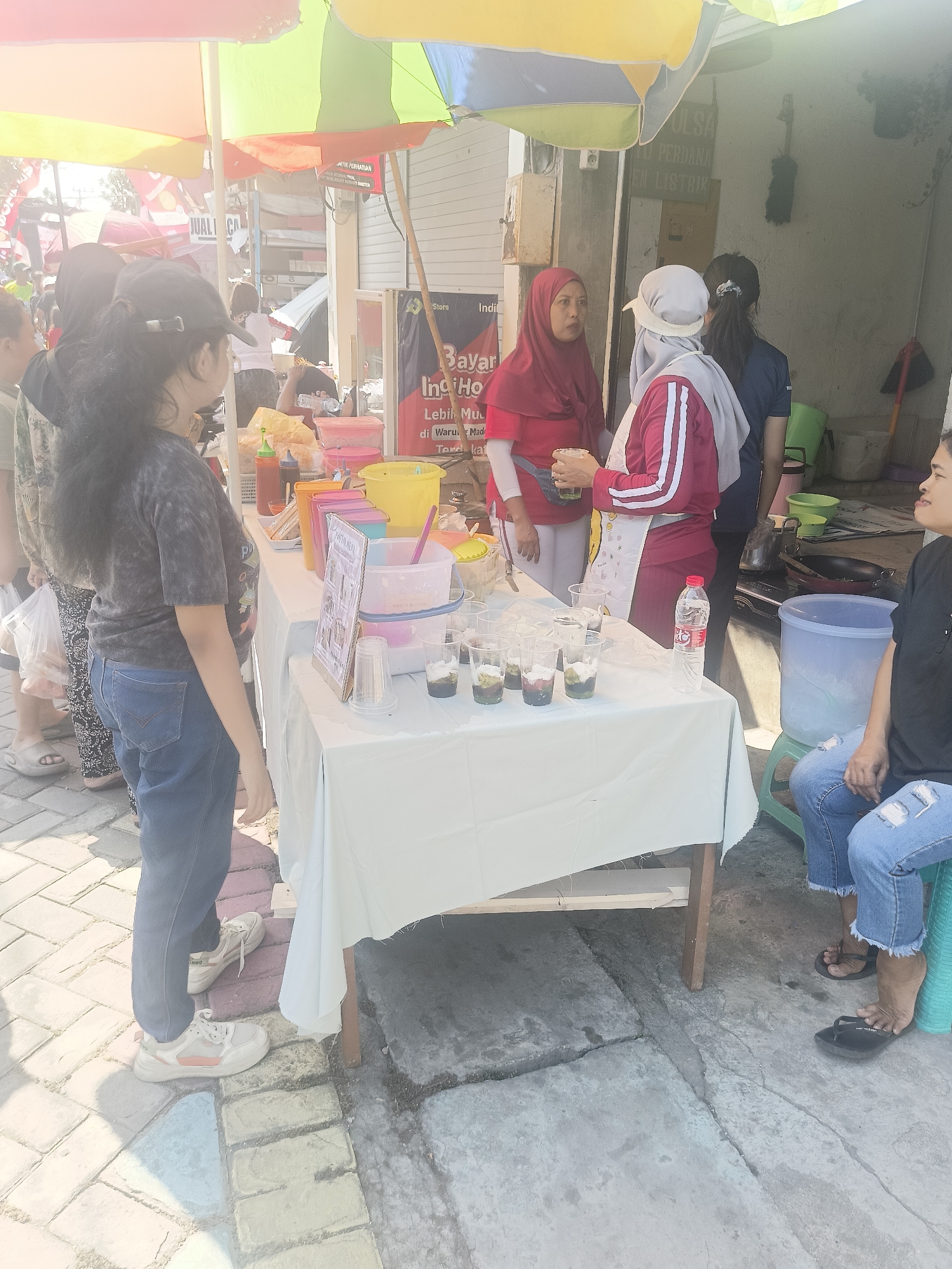 In Gedangan, Indonesia, on a sunny day, a street-level scene unfolds under colorful awnings. The focal point is a table draped in white, laden with various food and drink items. Several people are gathered around the table, suggesting a transactional or social interaction.
On the left, a young woman with long, dark hair, wearing a dark tie-dye t-shirt and blue jeans, stands facing the table, looking towards it. Her right hand is relaxed by her side, and her left arm is slightly bent. She is wearing white sneakers with orange accents.
In the center, two women are engaged in conversation. The woman on the left wears a red hijab and a red long-sleeved shirt, holding a clear cup with a green beverage. She has a darker green pouch or bag strapped across her chest. The woman next to her is wearing a white cap and a maroon jacket with white stripes on the sleeves. She is also holding a clear cup. Behind them, partially visible, is another person.
To the right of the table, a woman with dark hair, wearing a black t-shirt and ripped blue jeans, is seated on a green stool. She is looking to her left, away from the table, and is wearing black flip-flops.
The table itself displays an assortment of items. In the foreground, there are clear plastic cups containing layered desserts with green, brown, and white components, some with spoons. Behind these, several colorful plastic containers and bowls are visible, including pink, yellow, and orange ones, some holding food items that appear to be snacks or ingredients. A bottle of water with a red and white label stands upright on the table.
The environment suggests an outdoor marketplace or street vendor setting. The ground is paved with interlocking bricks in various colors, with some areas of concrete. In the background, buildings are visible, some with signage. One sign, partially obscured, has red lettering that appears to say "JUALI CA". Another sign in red text reads "Bayar Ind Ho" and below it "Lebih Mu... di Warung Mad...". The overall lighting indicates bright sunlight casting shadows.