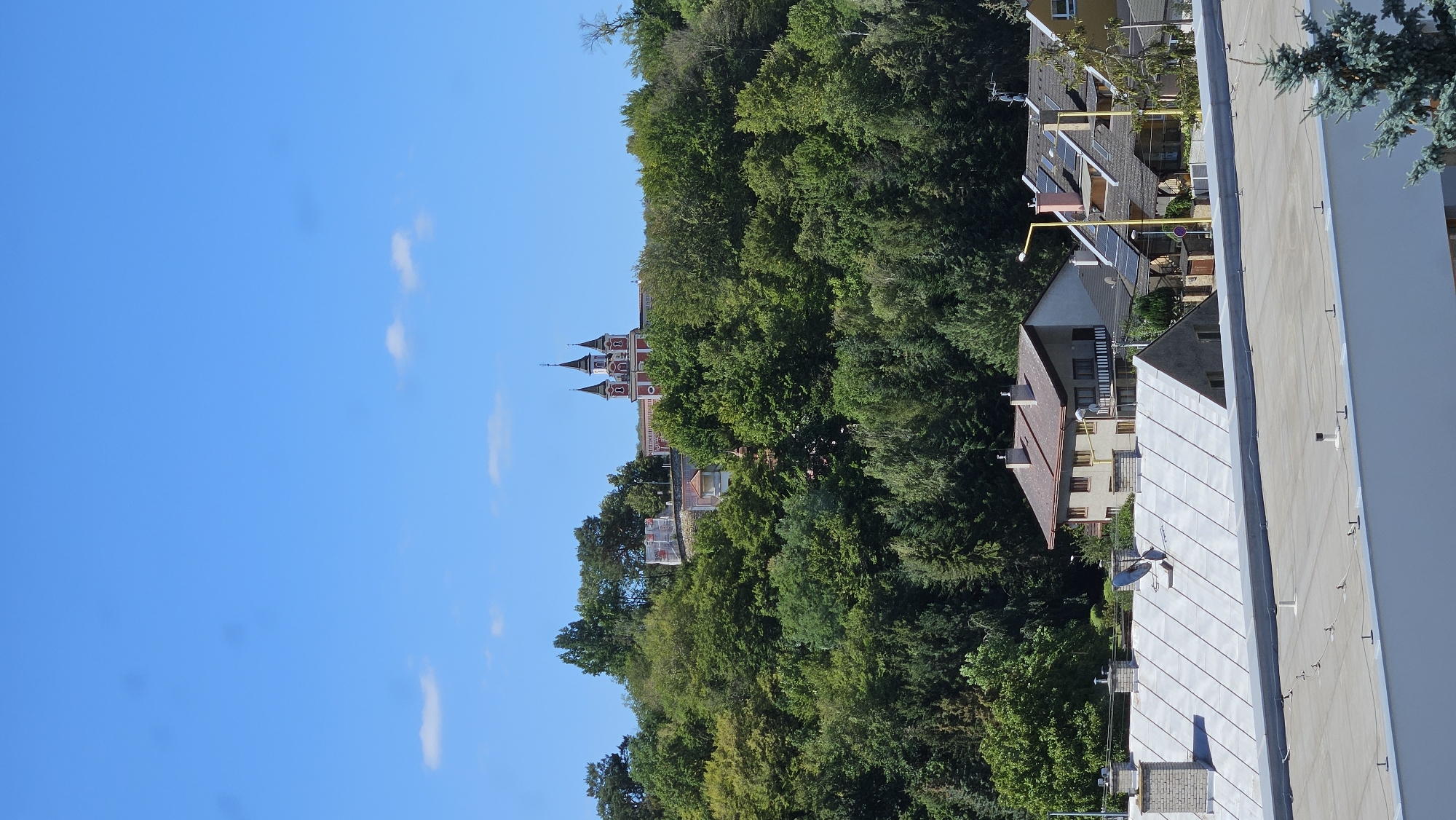 The image depicts a panoramic view of a hilly urban landscape under a clear blue sky with scattered white clouds. Dominating the upper portion of the frame is a red and white building with multiple spires, possibly a church or a monastery, situated on top of a densely wooded hill. Below the hill, a cluster of residential buildings with pitched roofs is visible. The foreground shows parts of several flat-roofed structures. The scene appears to be a daytime shot, likely during the summer, given the lush green foliage. While the exact location within Prešov, Slovakia is not specified by visual cues, the architectural style and the natural setting are consistent with areas found in and around Eastern Slovakian cities. There are no people or discernible human activity visible in the image.