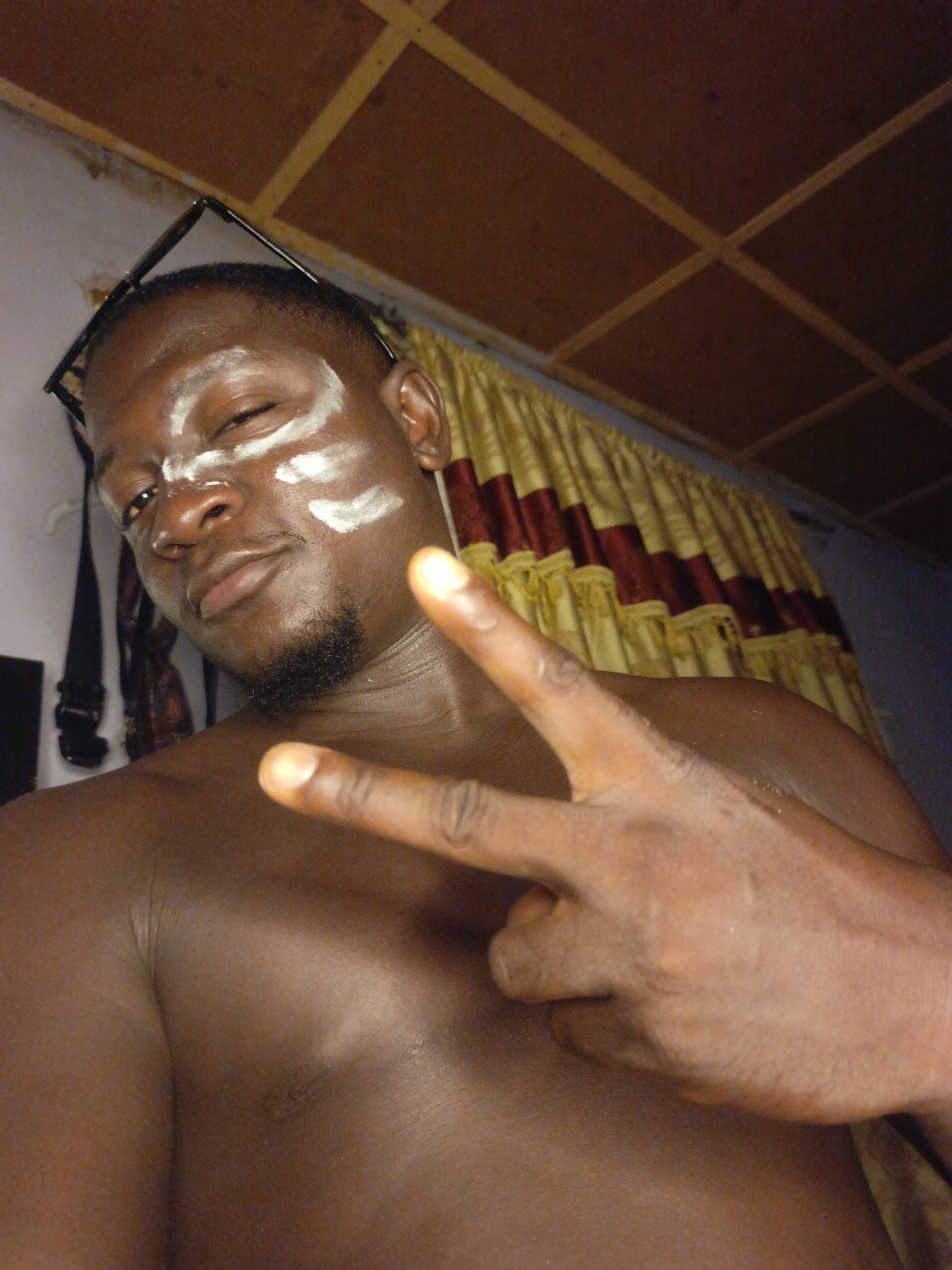 A topless, dark-skinned man is posing indoors, making a "peace" sign with his right hand. White markings are on his forehead and right cheek. Eyeglasses rest on top of his head. A curtain hangs in the background. The location is Akwanga, Nigeria.