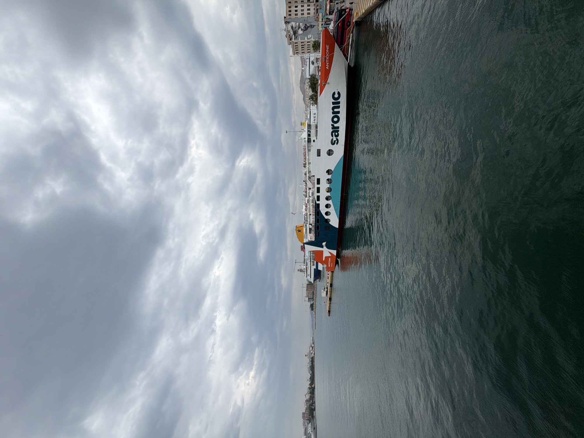 The image depicts a harbor scene in Piraeus, Greece, under a cloudy sky. A large ferry named "Saronic" is docked alongside a pier, with its orange and white hull prominently displayed. The water in the harbor is dark and rippled. Buildings and other ships are visible in the background, suggesting an urban port setting.