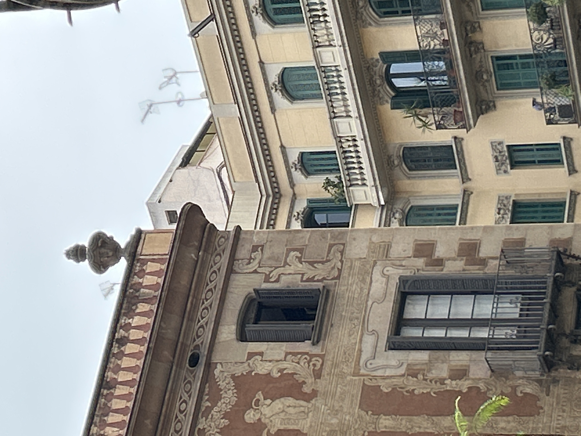 An upward-facing view captures the richly detailed facades of two buildings, likely in Barcelona, Spain, under an overcast sky. The image focuses on architectural ornamentation and fenestration, with no people visible.
The building in the foreground, positioned on the right side of the image (when oriented upright), exhibits a warm brown and beige facade. It is adorned with intricate painted or relief decorations, featuring swirling organic motifs, geometric patterns, and a discernible reclining figure (possibly a putto or cherub) within a larger floral-like design towards the lower portion. This building displays an arched window with dark, open louvered shutters, and a rectangular window fitted with dark frames and a decorative metal grate or small Juliet balcony. A sculptural finial crowns the upper right corner of this structure.
Behind and to the left of the foreground building, a second structure rises, characterized by a pale cream or light yellow facade. This building features multiple windows, all equipped with distinct dark green louvered shutters. Several balconies extend from its face, showcasing both intricate metal railings and classic stone balustrades. Some balconies host small potted plants, adding touches of greenery. The architectural style of both buildings appears consistent, suggesting a cohesive urban design, possibly Modernista.
The sky is a uniform, light grey, indicating an overcast or cloudy day. Faint, skeletal antenna-like structures are visible against the sky in the upper left background, suggesting rooftops or other distant structures. No discernible text is visible on either building.