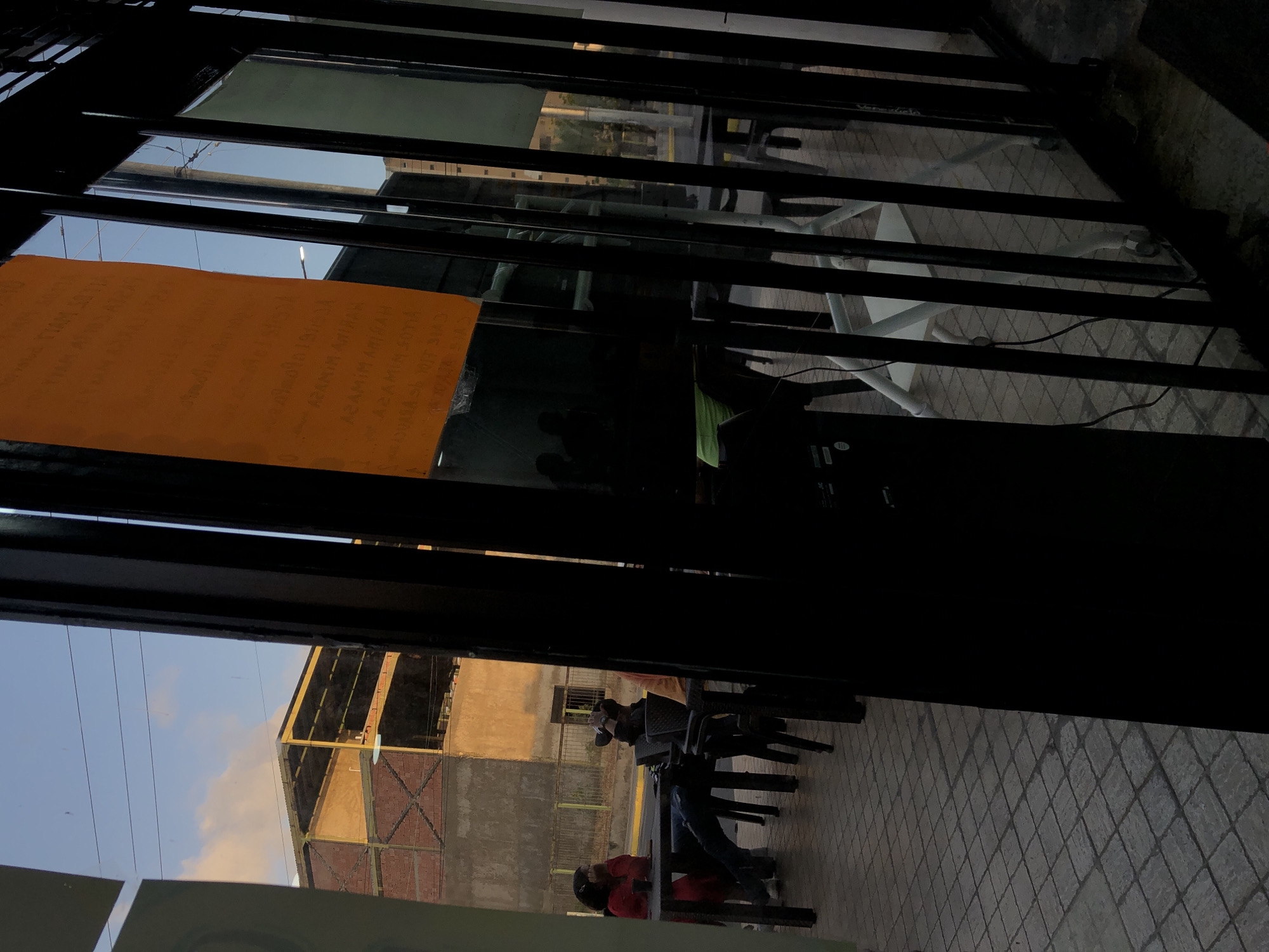 The image shows a view through a dark, gated doorway, likely an entrance to a business or establishment in Juan Griego, Venezuela. An orange sign is taped to the gate. Outside, a paved area with a diamond pattern is visible. Two people are sitting at a table near a building with a mix of tan and gray walls. The sky is a pale blue.