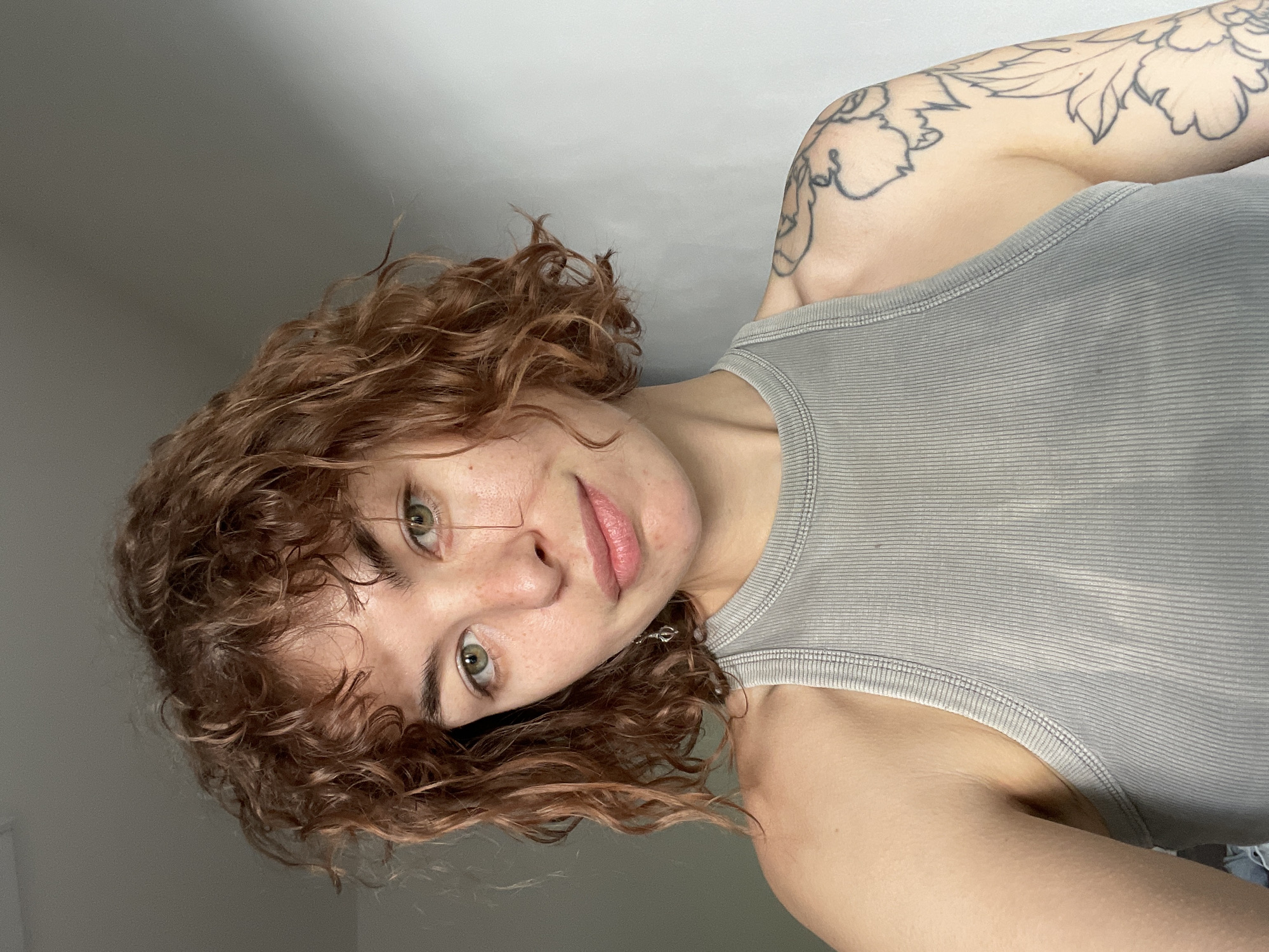 A person with curly reddish-brown hair, green eyes, and fair skin is visible from the chest up, wearing a ribbed grey tank top. The person's left arm is visible, featuring a black-ink floral tattoo. The background is a plain, light grey wall. The image appears to be taken at an angle, tilting to the right. No information confirms the location is Boreti, Montenegro.