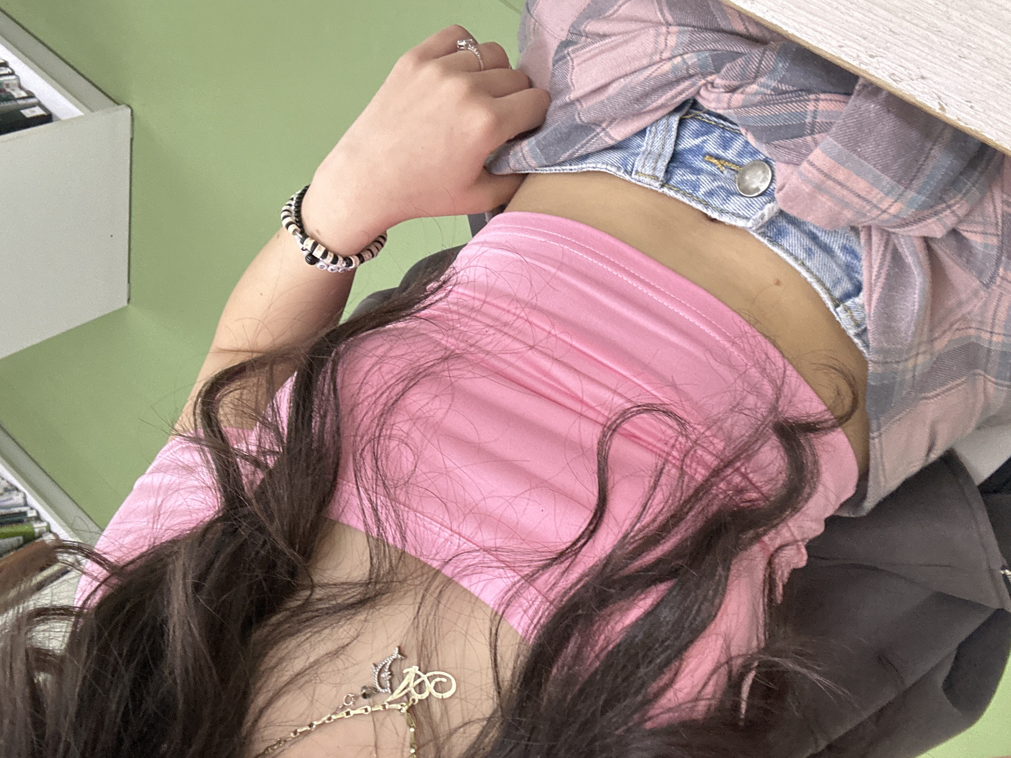 The image shows a person wearing a pink top, denim shorts with a button, and a pink and grey plaid shirt. They are also wearing a bracelet and a necklace. The background is a green wall and a white shelf unit. The location is unknown.