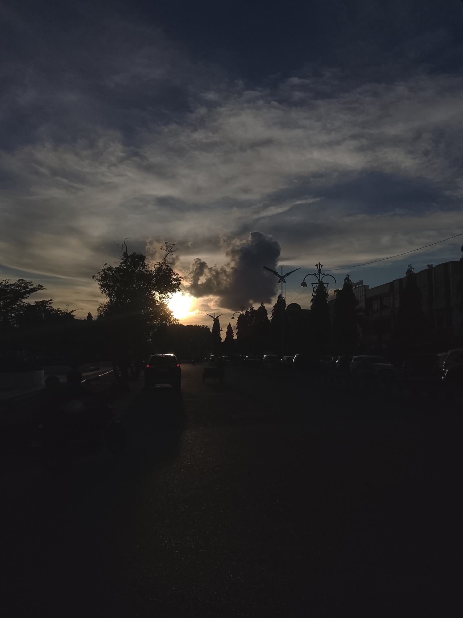 The image depicts a street scene in Lhokseumawe, Indonesia, during what appears to be sunset or dusk. Silhouettes of trees, buildings, and vehicles are visible against a dramatic sky with dark clouds and a bright sun peeking through. Several cars are on the road, and some individuals are seated or standing on the side. The street itself is paved and appears wet, reflecting some of the ambient light. Lampposts and power lines are also discernible. The overall impression is one of an urban environment transitioning from day to night, with a focus on the atmospheric conditions.