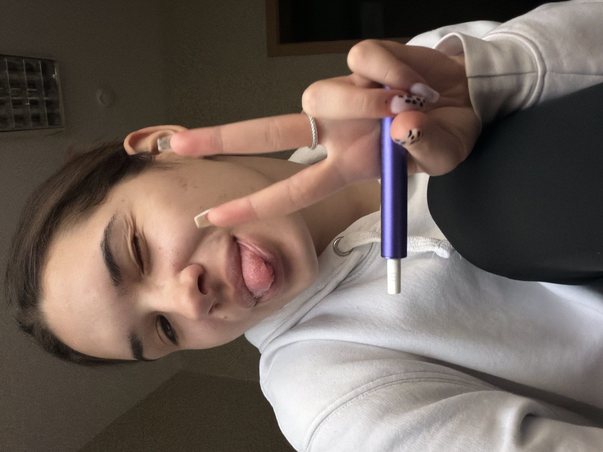 A close-up, rotated selfie features a young individual, likely female, captured indoors. The subject has dark brown hair, pronounced eyebrows, and fair skin, and is shown mid-expression with one eye winking and their tongue playfully sticking out. They are wearing a white hooded sweatshirt with visible drawstrings and stitching details.
One hand is prominently displayed, making a "V" sign with the index and middle fingers extended. The fingernails have a light, possibly pink or nude, base coat with dark, speckled patterns, reminiscent of an animal print like cow print. A silver-colored, twisted or braided ring is worn on the index finger. Between the index and middle fingers, the person holds a slender, purple cylindrical device with a white tip, strongly resembling a vape pen.
The background is dimly lit and indistinct, suggesting an indoor setting with low ambient light. In the upper left background, a rectangular metallic vent or grate is visible against a dark wall. Hints of other dark furniture or objects are present in the deeper background. The overall mood conveyed is casual and playful. There is no visible text within the image. The context places this scene in Sofia, Bulgaria.