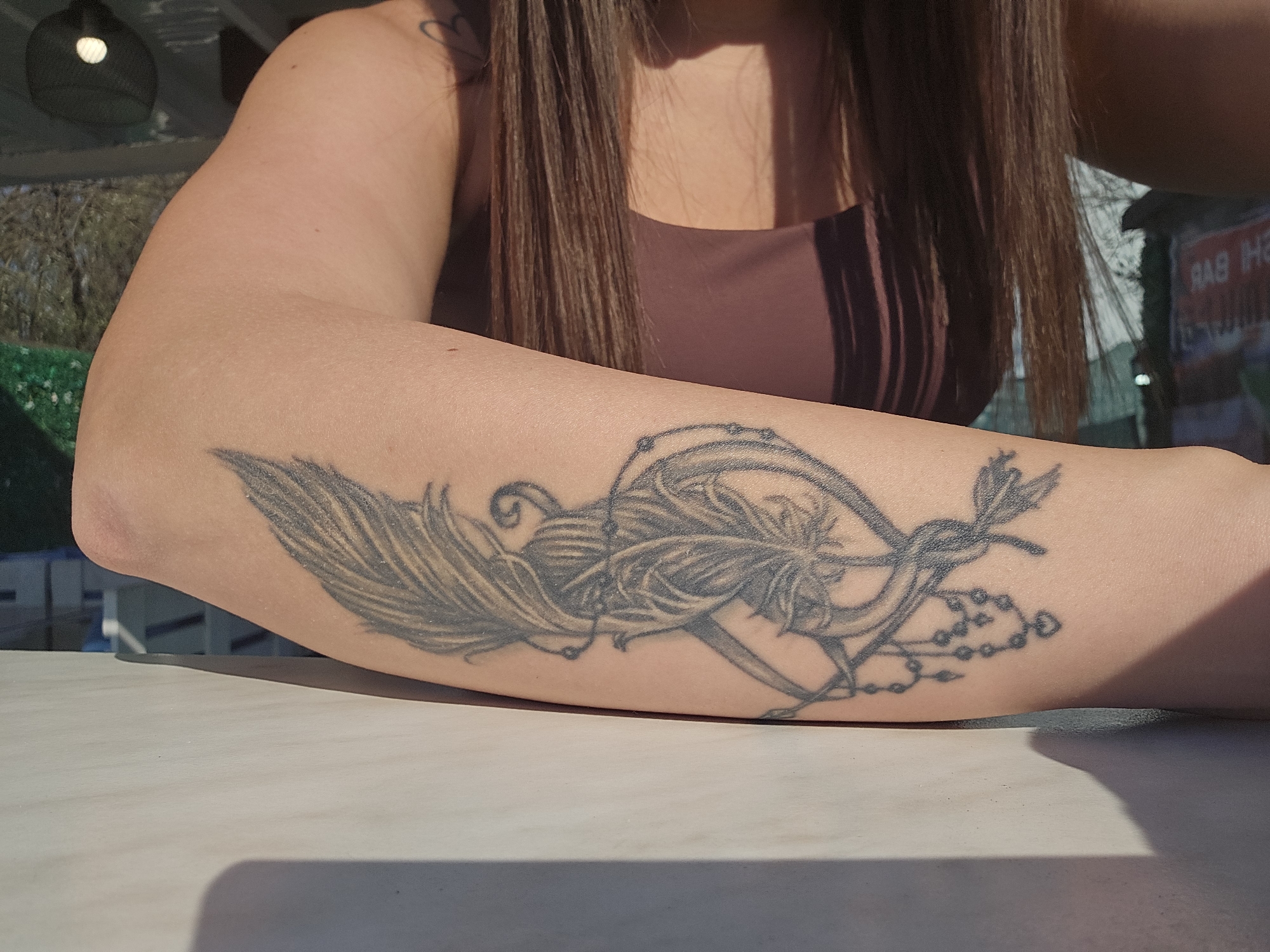 A woman's forearm rests on a white, possibly sunlit, table. The arm has a large, black and gray tattoo featuring a feather, an infinity symbol, and beads. The woman is wearing a tan top. Her brown hair hangs down. In the background are hints of foliage and an out-of-focus sign written in a language that appears to be Bulgarian, suggesting a location in Burgas, Bulgaria. Other elements are blurry, obscuring the exact setting.