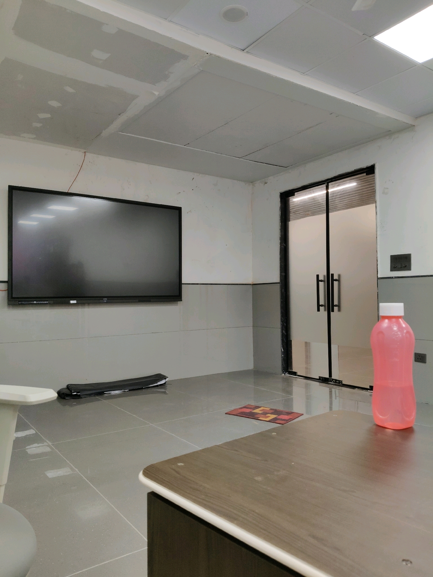 This image depicts an interior scene, likely an office or conference room, in Goner, India. The room appears to be under construction or renovation, with exposed ceiling panels and unfinished walls. A large, dark-colored screen is mounted on the wall. In the foreground, a light brown desk surface is visible. To the right, a pink plastic bottle is placed on the desk. A set of glass doors with black handles leads to another area of the building. The floor is tiled with a light grey material. A black object, possibly a rolled-up mat or document case, lies on the floor near the wall.