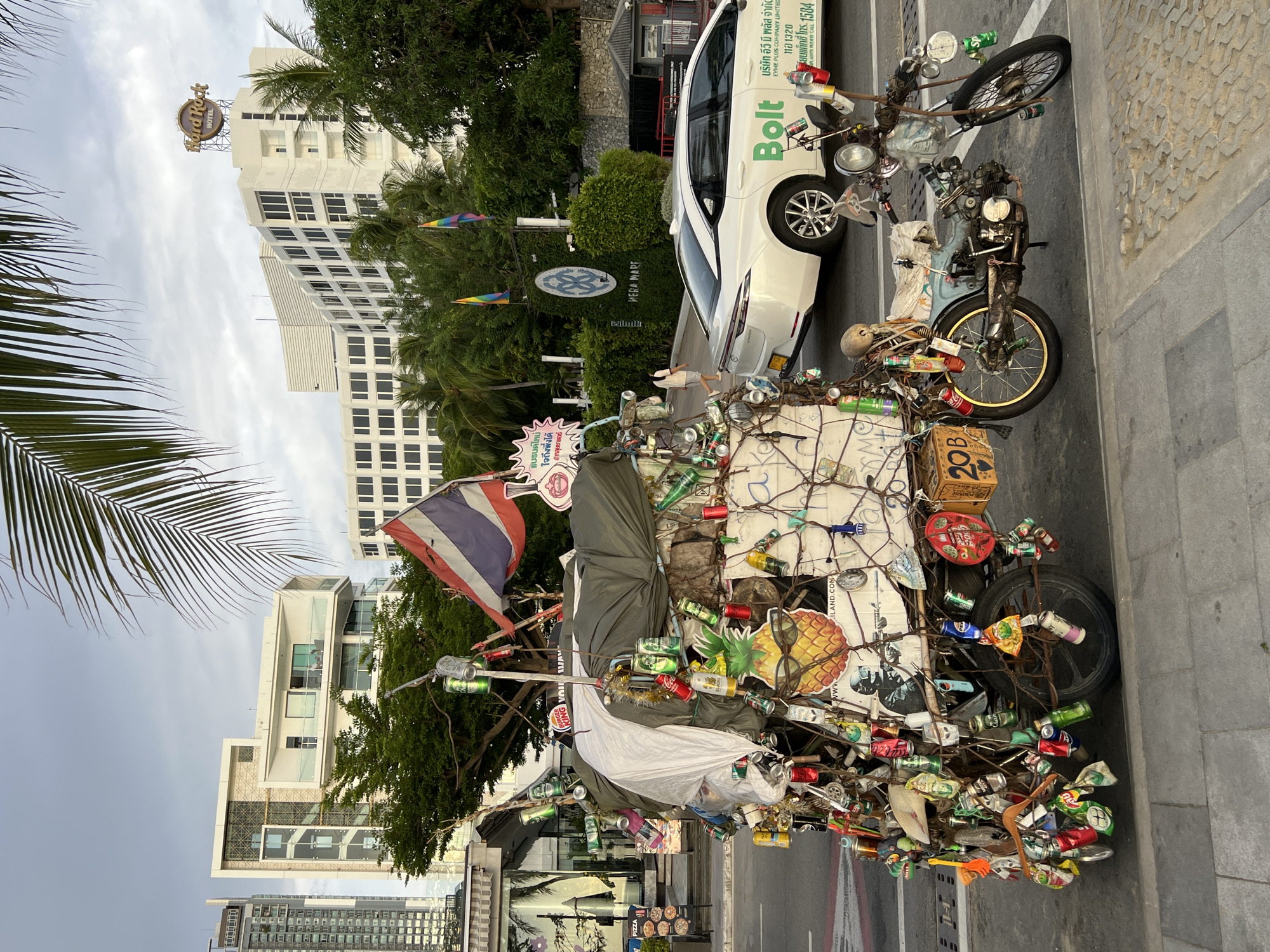 The image depicts a heavily decorated motorcycle and sidecar laden with numerous empty beverage cans, bottles, and other assorted items, parked on a sidewalk in Pattaya City, Thailand. The motorcycle is a vintage model, painted blue and silver, with a weathered appearance. It is attached to a two-wheeled sidecar, which is almost entirely obscured by a dense arrangement of hanging objects, including beer cans, plastic bottles, and what appear to be small toys or ornaments. A Thai flag is prominently displayed on a pole above the sidecar.
In the background, modern buildings suggest an urban environment. A tall, slender high-rise building is visible on the left, followed by a white building with balconies and contemporary architectural features. Further to the right, a larger white building with the "Hard Rock Hotel" signage on its roof is discernible, indicating a tourist or entertainment district. A white Bolt ride-sharing vehicle is parked in the street to the right of the decorated motorcycle. A woman, appearing to be a pedestrian, walks in the middle ground near a building with the "HERA MARE" sign. Lush green trees and palm fronds frame the scene, suggesting a tropical or subtropical climate. The sky is overcast, with a soft, diffused light. The sidewalk is paved with grey tiles, and a street with traffic markings is visible in front of the sidewalk.