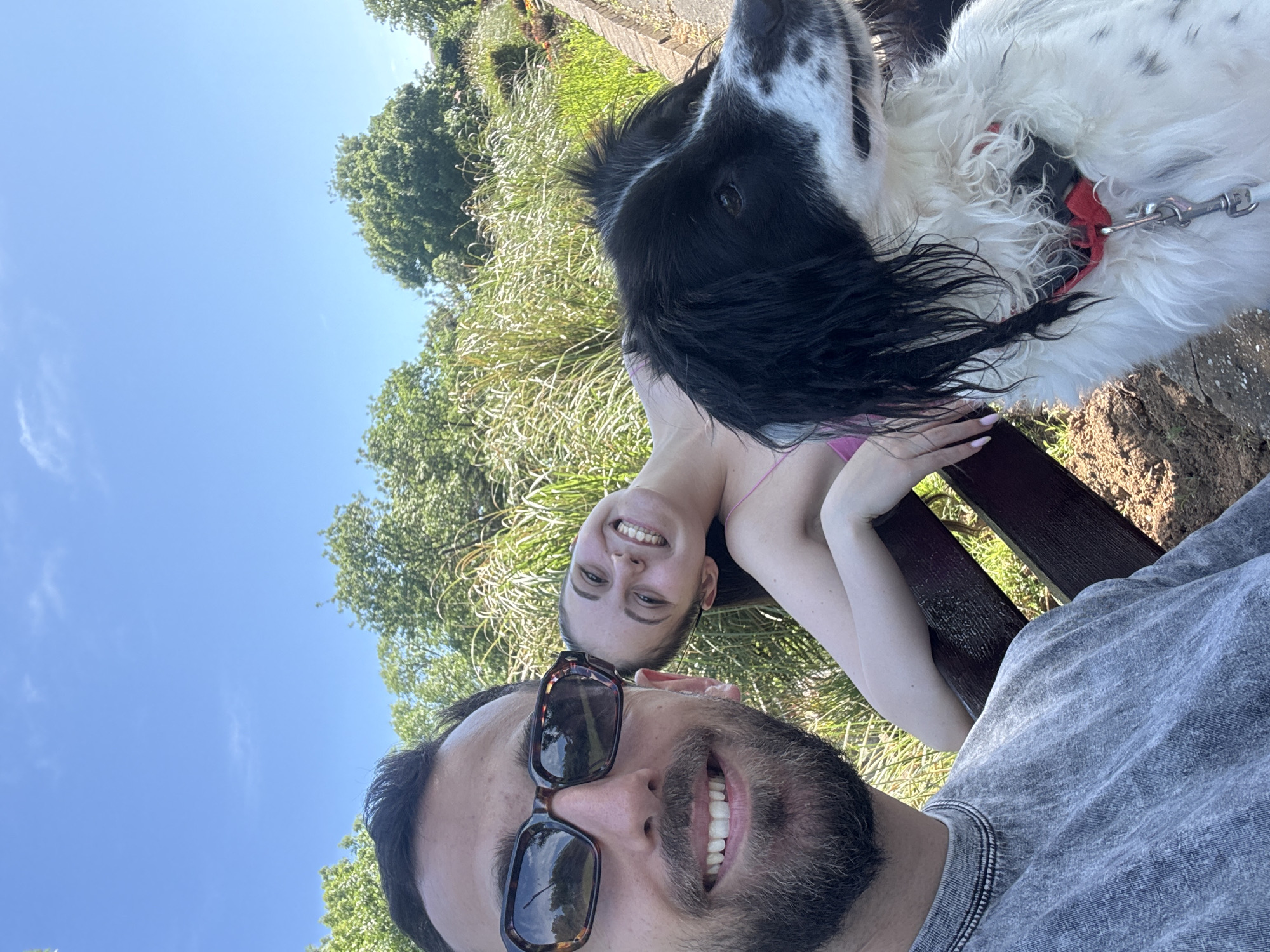 A selfie captures a man with a beard and sunglasses in the foreground, looking up and smiling. Behind him, a woman smiles towards the camera, leaning on a wooden bench. A black and white dog with a red collar is prominently positioned above them. The background reveals trees, vegetation, and a clear blue sky. The location is Tsarevo, Bulgaria.
