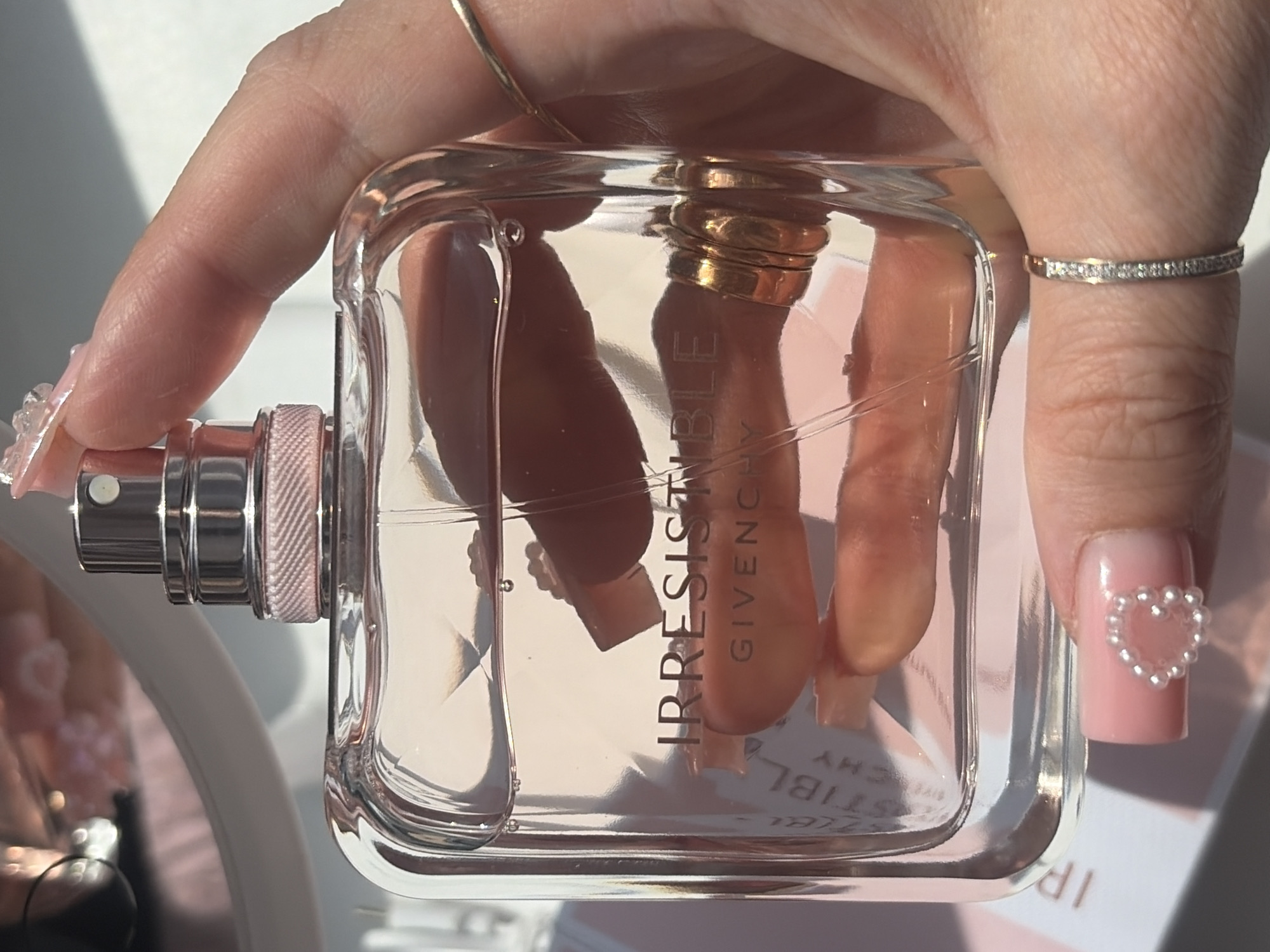 A close-up image captures a right hand holding a clear glass perfume bottle, set in the city of Novosaratovka, Russia. The rectangular bottle, with rounded edges, contains a pale pink liquid with visible air bubbles. The text "IRRESISTIBLE" and "GIVENCHY" is printed in dark lettering on its front surface. A silver metallic spray mechanism with a textured light pink band is affixed to the top, and a clear spray tube extends into the liquid. The hand, with light skin tone, displays well-manicured nails; the thumbnail features a pink and white ombre design adorned with a white pearl heart-shaped embellishment. The index finger wears a thin, plain gold-colored ring. The middle finger is adorned with three thin, plain gold-colored rings, and the thumb bears a thin gold-colored ring with small, clear stones. The background is softly blurred, showing light-toned surfaces and reflections, including parts of a mirror or shiny object in the upper left and a partially visible pinkish-white box with "IRRESISTIBLE" text at the lower left.