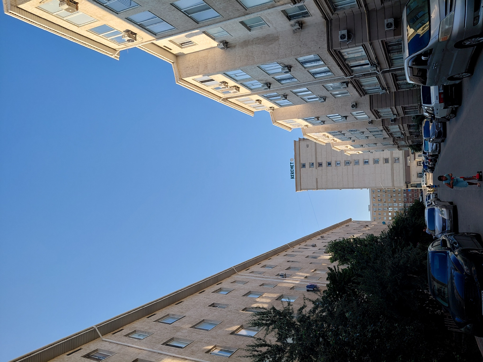 In the city of Aktau, Kazakhstan, a street view is captured on a bright, clear day, with a vast blue sky dominating the upper half of the frame. Two multi-story residential buildings, characterized by their light-colored facades and numerous windows, line either side of the street, creating a canyon-like effect. The building on the left appears to be an older style apartment block, while the building on the right showcases more modern architectural elements, including visible air conditioning units mounted on its exterior.
The street is lined with parked cars, suggesting a residential area. A black sedan is parked prominently in the foreground on the left, followed by a row of other vehicles, including a silver minivan and several cars. In the mid-ground, a young child, possibly a girl in a blue dress, is riding a red scooter. Further down the street, more buildings are visible, including a taller, lighter-colored structure with the word "KEREMET" and the number "49" visible on its upper facade. The environment appears to be a typical urban setting in Kazakhstan.