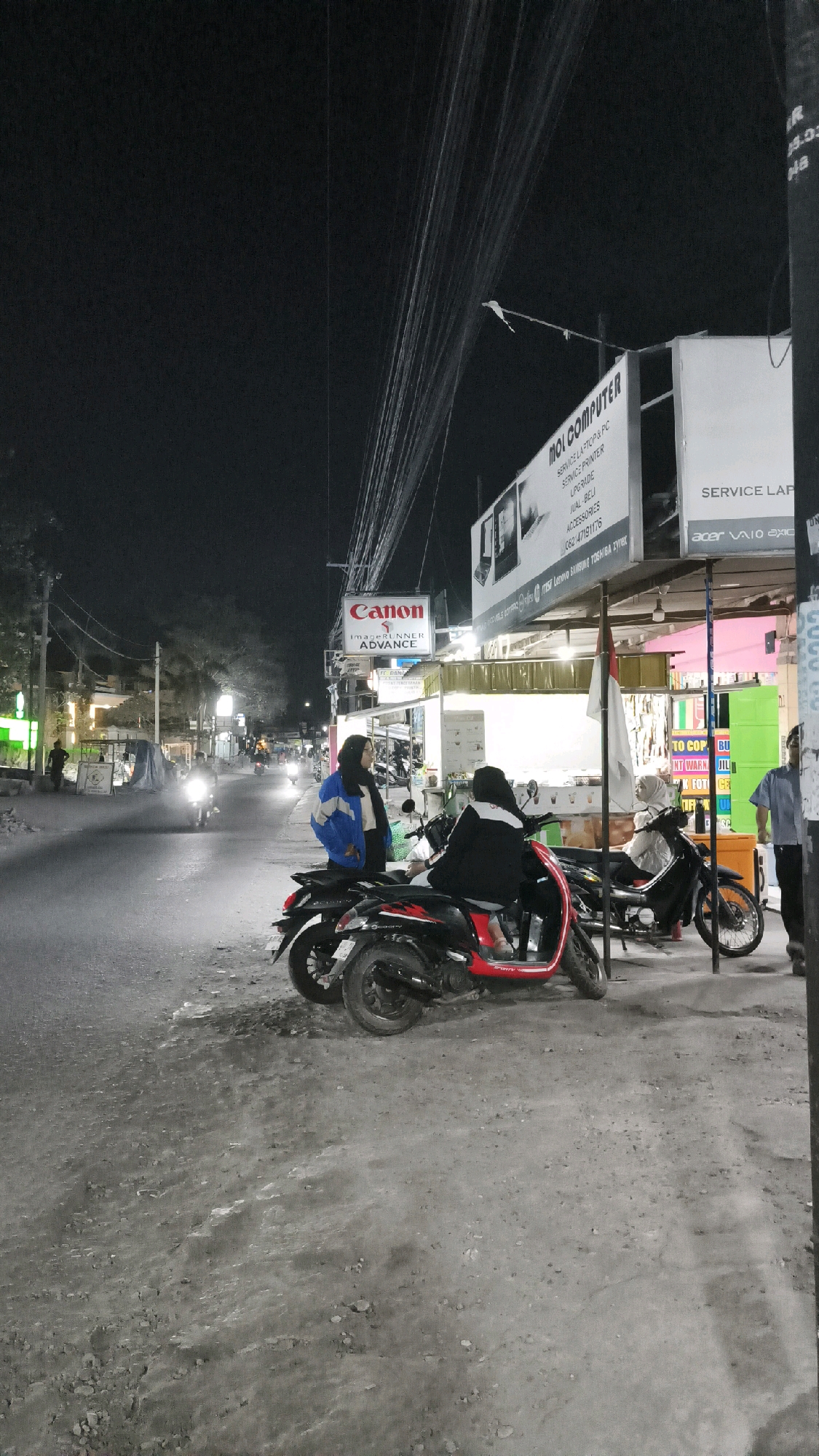 At night, on the side of a paved road in the city of Kartasura, Indonesia, two young women are sitting on a red scooter. The woman in front is wearing a black jacket and the woman behind her is wearing a white jacket with black trim. Several other scooters are parked on the side of the road. A sign for "MOL COMPUTER" is visible, along with other signage for "Canon" and "Service Laptop & PC". The street is illuminated by streetlights and the lights from the shops. There is a general sense of activity on the street, with other people and vehicles visible in the distance.