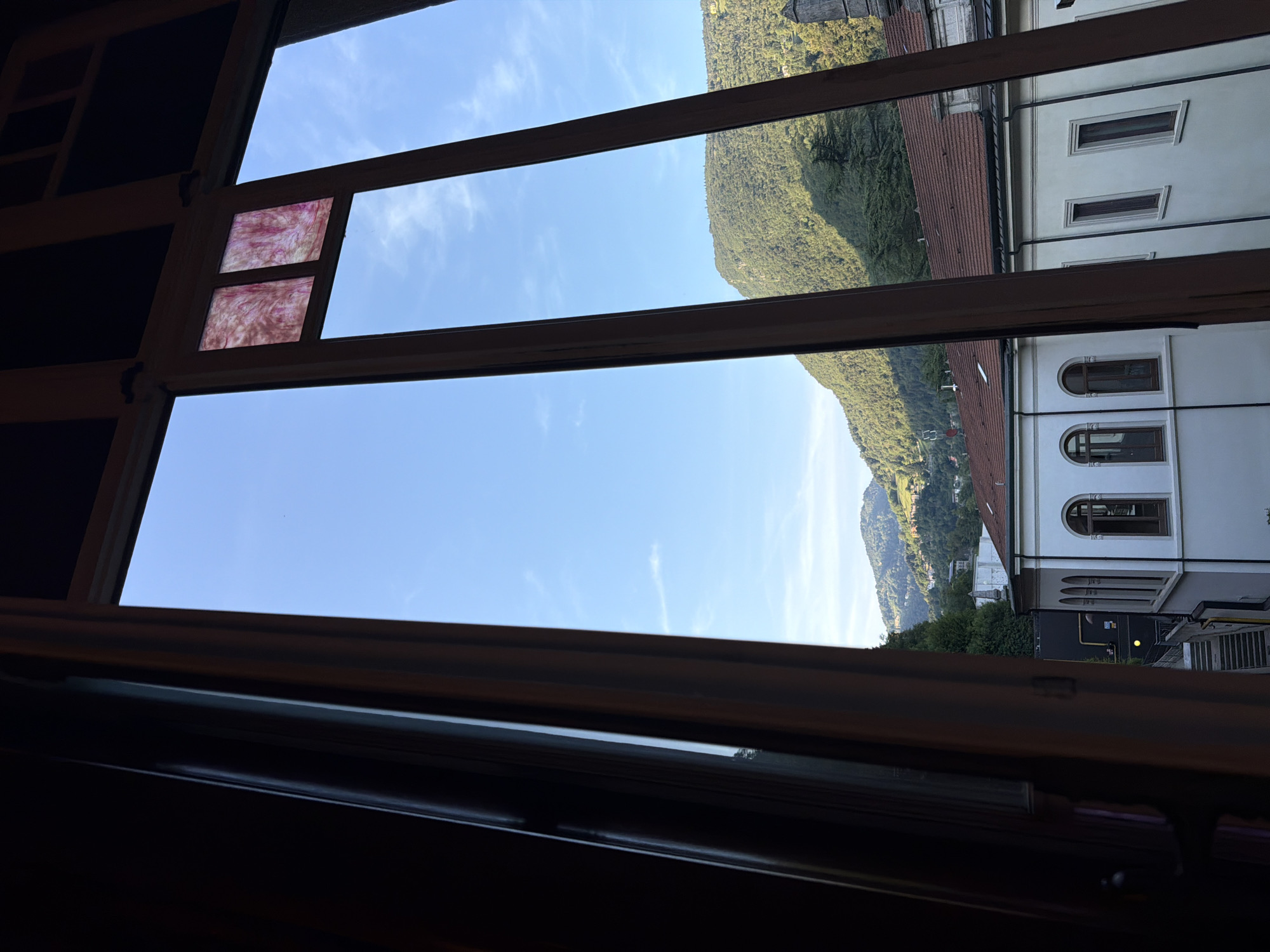 This media file depicts a view from a window in San Pellegrino Terme, Italy. The scene is dominated by a clear blue sky with sparse white clouds, suggesting fair weather during the daytime. The window frame, made of dark wood, divides the view into several sections.
In the upper left portion of the window, a small pane of decorative stained glass with swirling pink and white patterns is visible. The main view through the window showcases a lush green mountainside, characteristic of the Italian Alps, dotted with trees and some buildings nestled into the slope. The lower part of the view reveals buildings with white facades and tiled roofs. One building features arched windows, typical of older European architecture. A yellow circular object, possibly a light fixture or sign, is visible on one of the buildings. The overall impression is a peaceful, scenic landscape bathed in natural light.
No people or specific activities are discernible in the image. The composition, looking outwards from an interior space, evokes a sense of quiet observation or contemplation. There is no visible text in the image.