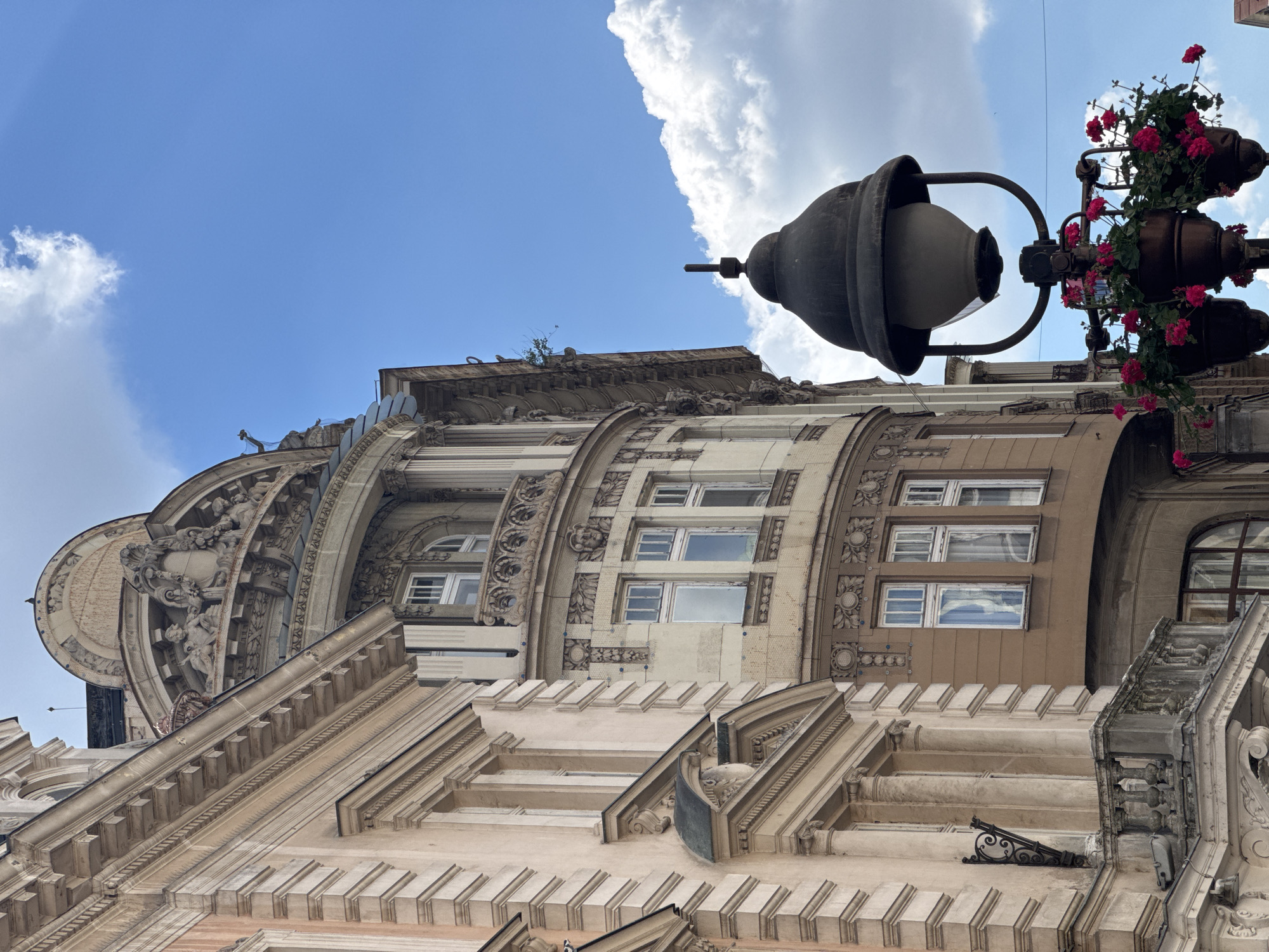 This is a low-angle shot of ornate architecture and a street lamp against a bright blue sky with scattered clouds, likely in Belgrade, Serbia. The buildings feature classical architectural elements, including columns, detailed carvings, and curved facades. The street lamp is dark metal, with a large, domed lantern and three hanging planters filled with vibrant pink flowers. The overall impression is one of historical European urban scenery.