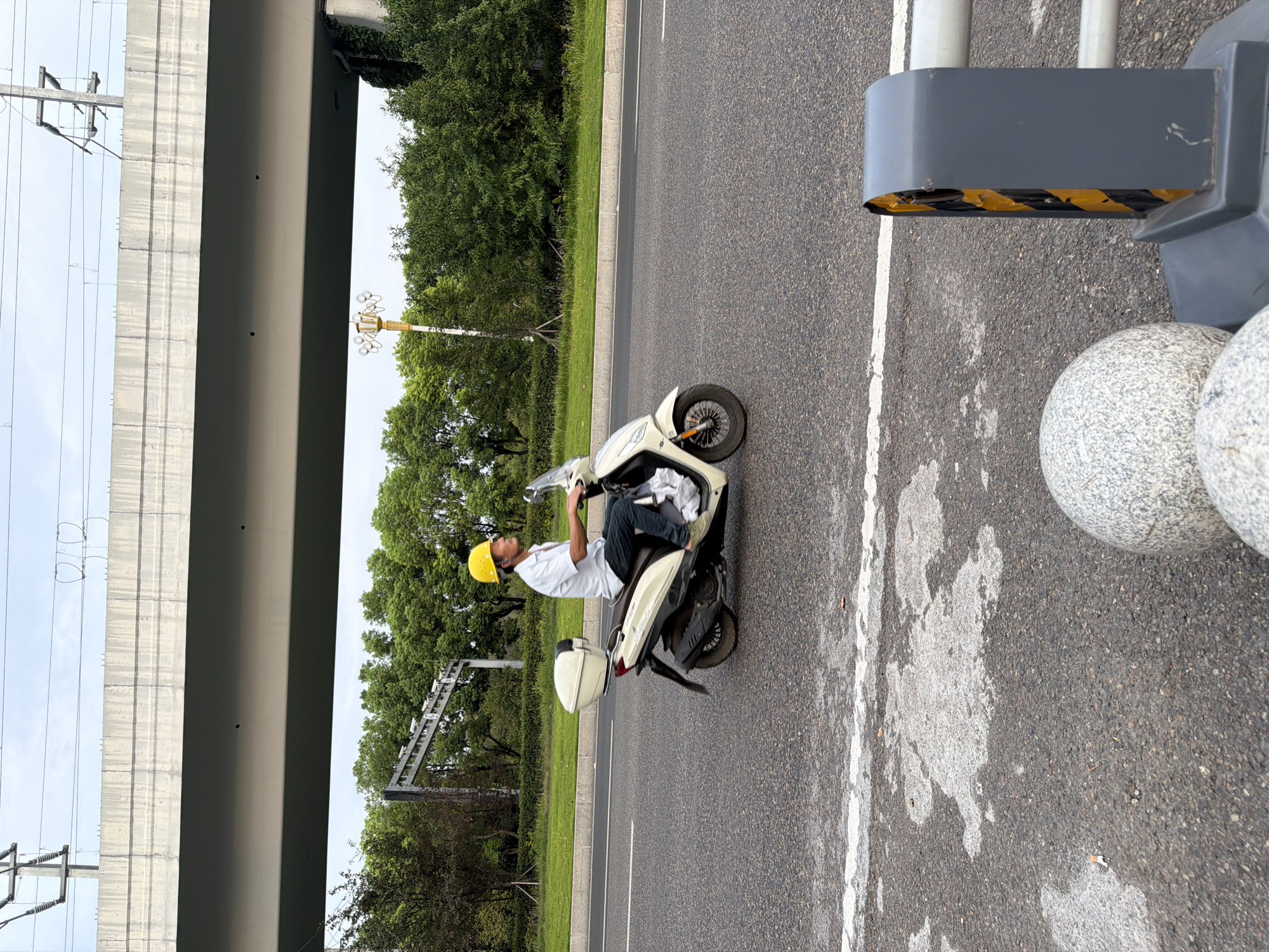 A man in a yellow hard hat rides a cream-colored scooter on a paved road. He wears a short-sleeved white shirt and dark pants. The scooter has a white storage box on the back. The road is lined with green grass and trees. In the background, a large concrete overpass crosses above the road, with power lines running overhead. The sky is overcast. The scene appears to be in the city of Jiangjin, China. In the foreground, to the right, is a dark grey barrier with yellow and black reflective tape, and two decorative stone spheres.