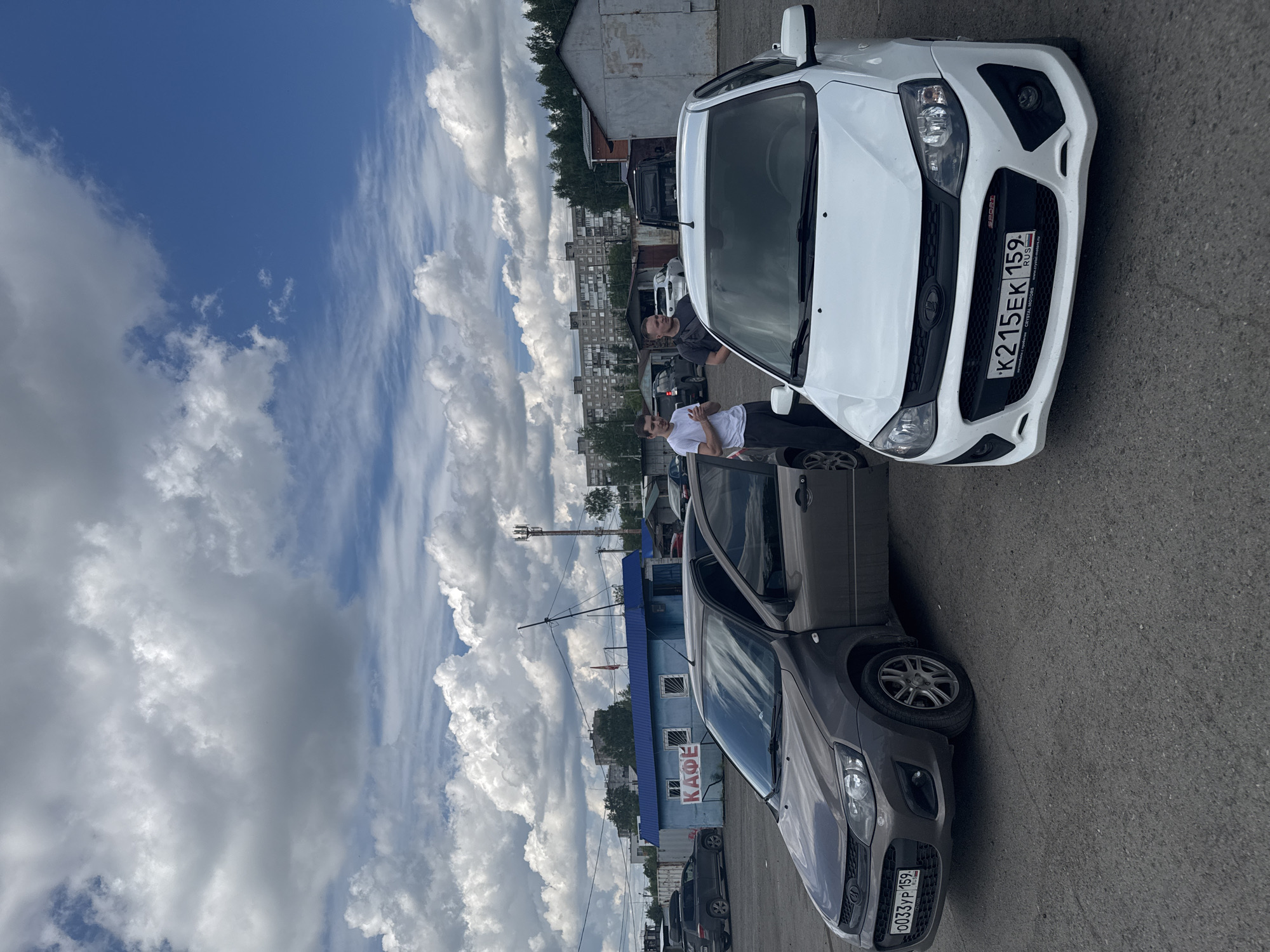 The scene depicts two cars parked on an asphalt surface under a cloudy sky. A white Lada Granta with license plate K215EK 159 RUS is parked facing the camera. A gray Lada Granta with license plate O033YP 159 RUS is parked beside it. Two men are standing near the cars. One man is wearing a white t-shirt. In the background, there are buildings, trees, and a blue structure with the Russian word "КАФЕ" (cafe). Based on the license plates, the location is likely Perm, Russia.