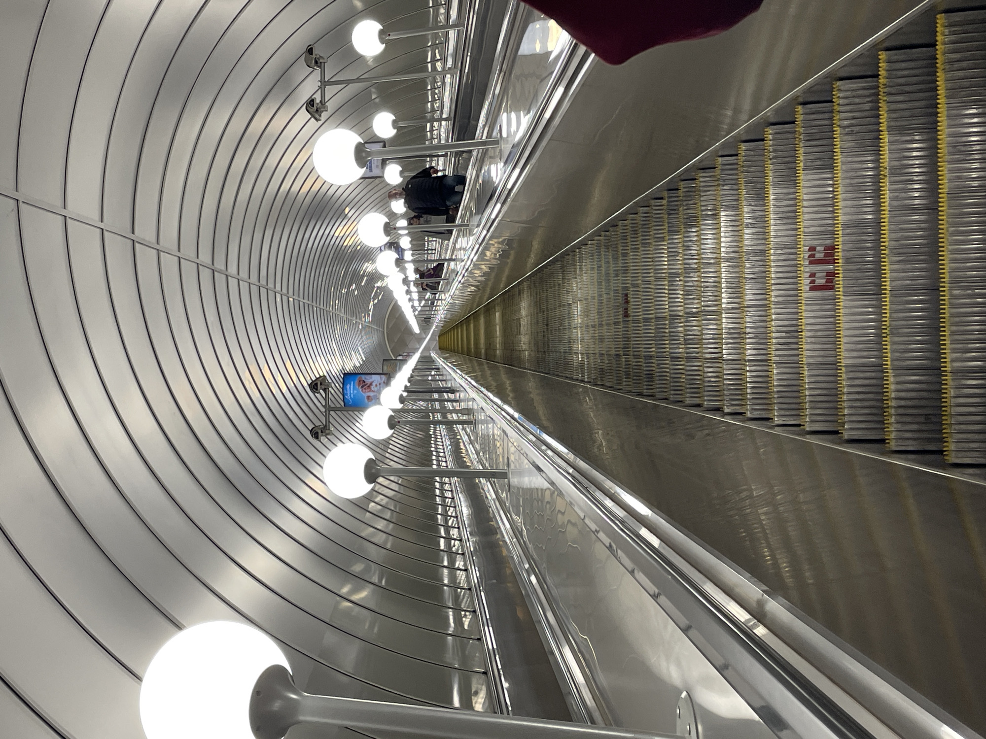 The photograph depicts a downward-angled view of an escalator inside a subway station. The walls are lined with curved, metallic panels that converge in the distance, creating a tunnel-like effect. A row of spherical lights is mounted along one side of the wall. People can be seen riding the escalator in the center. There is an advertisement on the wall. Based on the architectural style and details, this scene is most likely at the Avtovo metro station in Saint Petersburg, Russia.