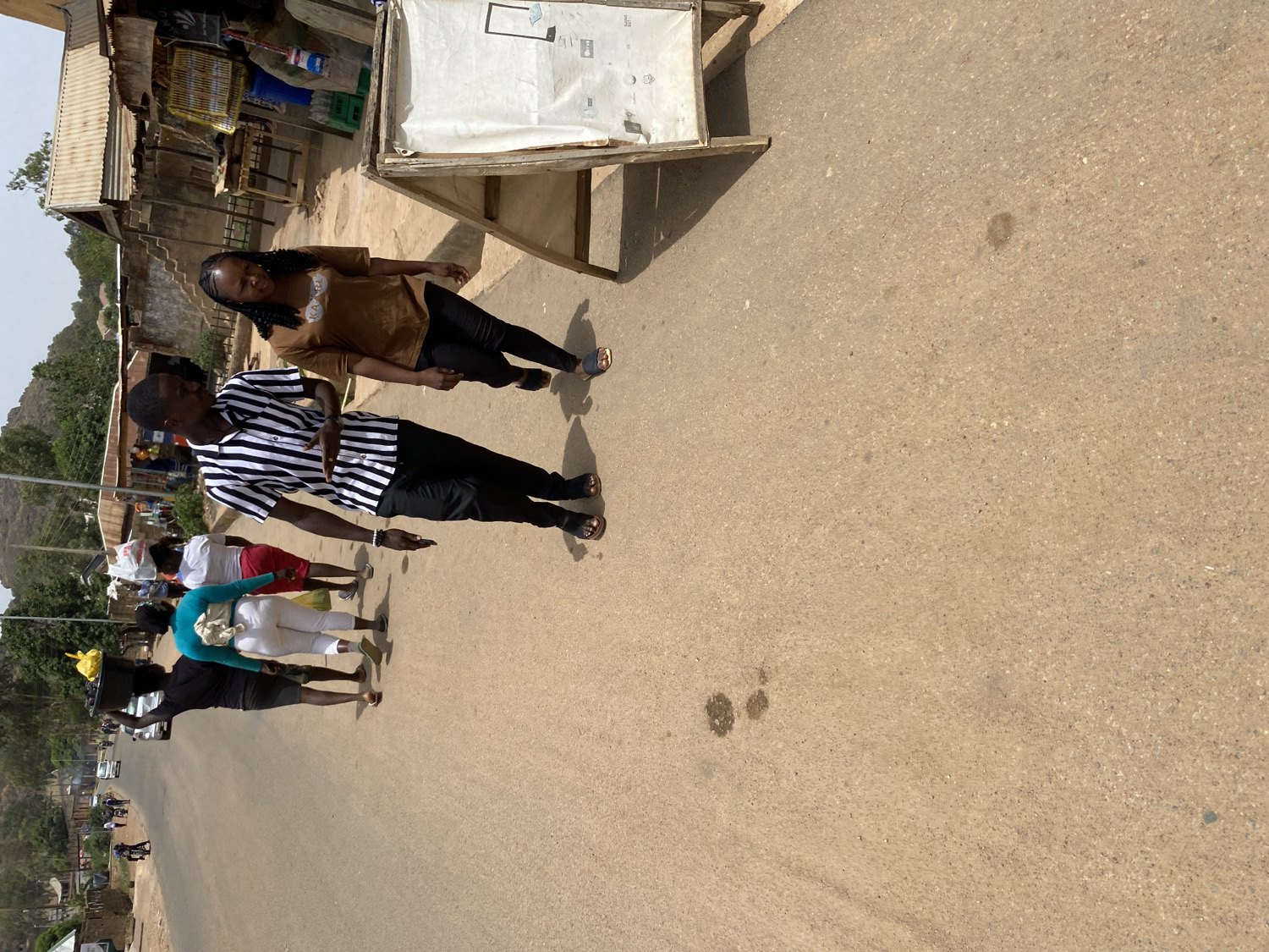 The image captures a vibrant street scene, likely in a market or commercial area, under bright daylight with clear skies. The setting is consistent with the provided location of Jos, Nigeria.
In the foreground, two main figures are prominent. A man on the left wears a black and white striped short-sleeved shirt, black trousers, and dark sandals, looking towards the right. Beside and slightly behind him is a woman with long, dark braided hair, wearing a brown short-sleeved top, black trousers, and light-colored sandals. She is looking to the left and has a faint smile.
Further down the street, several other people are present, indicating a busy environment. One individual, dressed in a dark top and shorts, is seen carrying a yellow basin on their head, a common practice for transporting goods. Other people are casually dressed in various attire, including a light blue top with white pants and a white top with red shorts.
On the left side of the road, there are structures resembling market stalls or small shops with corrugated iron roofs, suggesting commercial activity. On the right side, a wooden stand with a white covering is visible, on which some indecipherable black text and a rectangular outline can be seen. The road itself is a light brown-grey paved surface, showing a few dark stains. In the distant background, hills or mountains are discernible on the left, and a vehicle is partially visible further along the road. The overall scene depicts typical daily life and movement within a community.